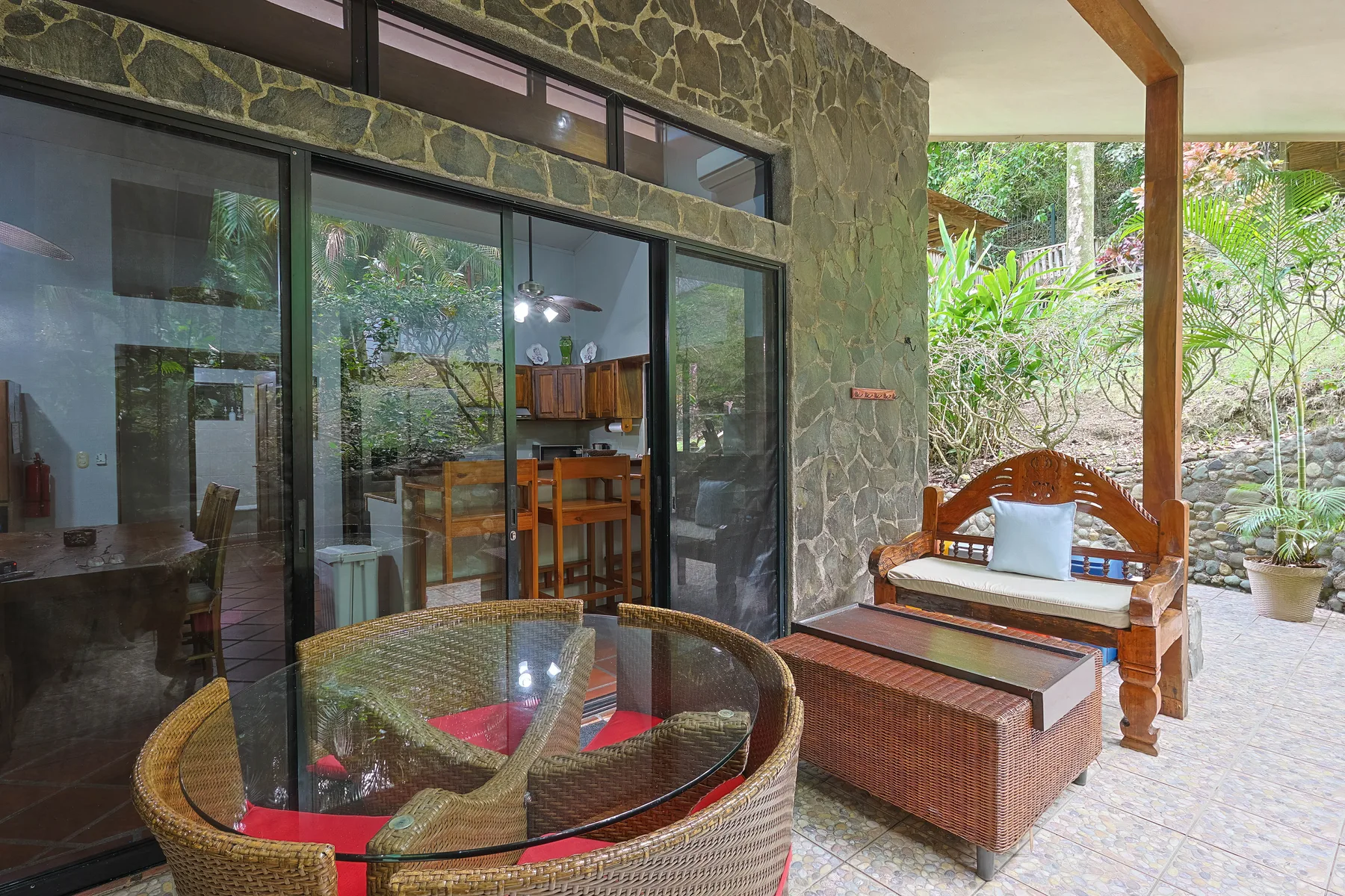 Un patio cubierto con asientos de mimbre y una mesa de cristal da a un jardín frondoso.