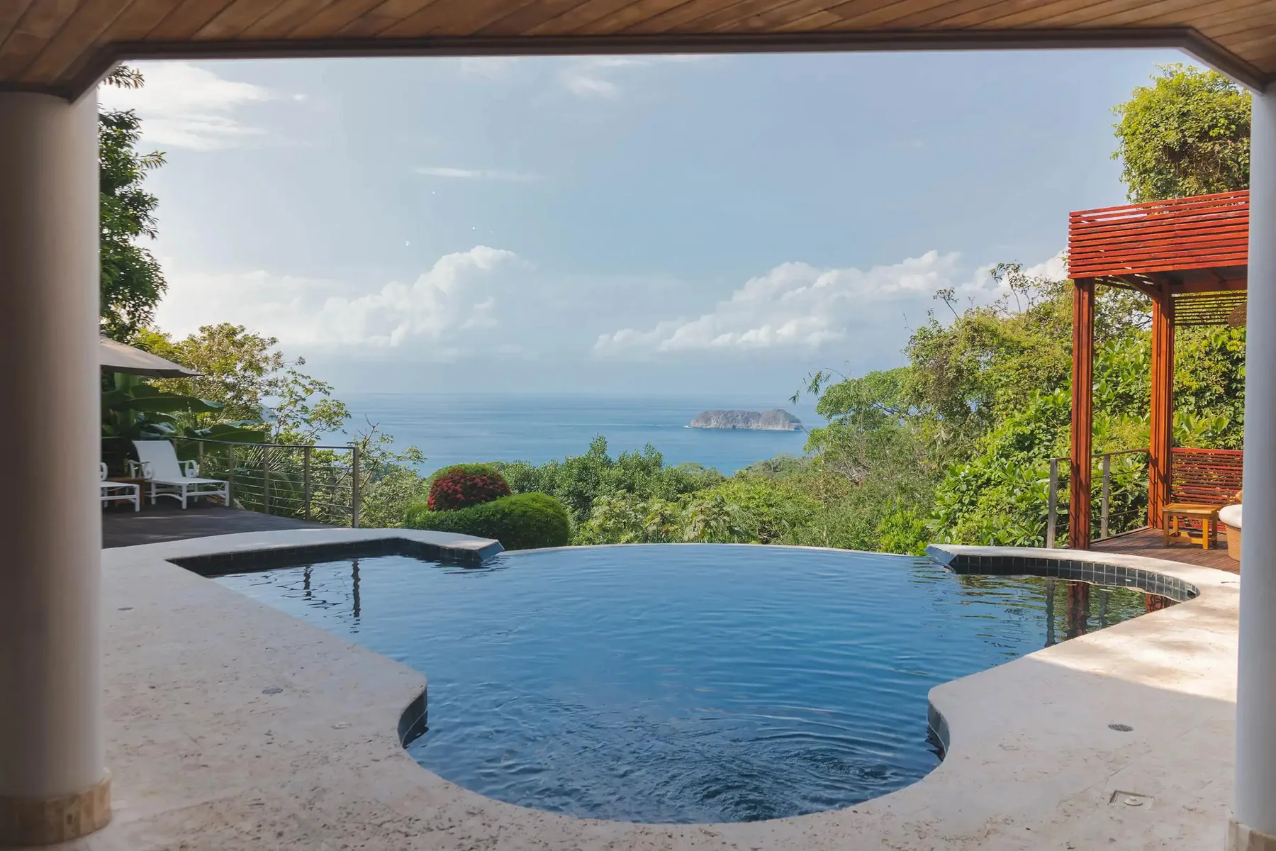 An outdoor pool overlooks the ocean, with greenery and a shaded seating area nearby.