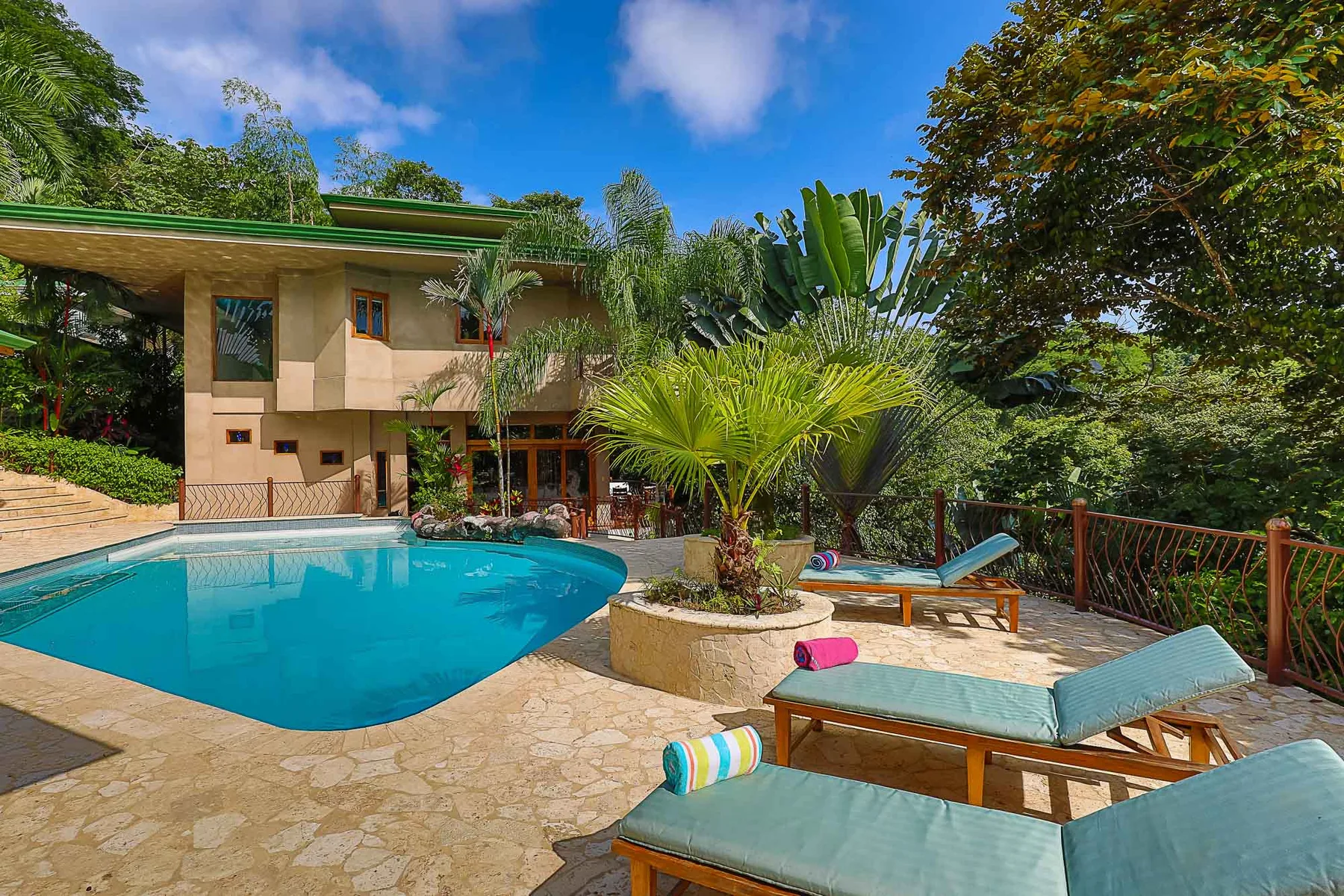 A tropical outdoor pool beside a house, with lounge seating and surrounding greenery.