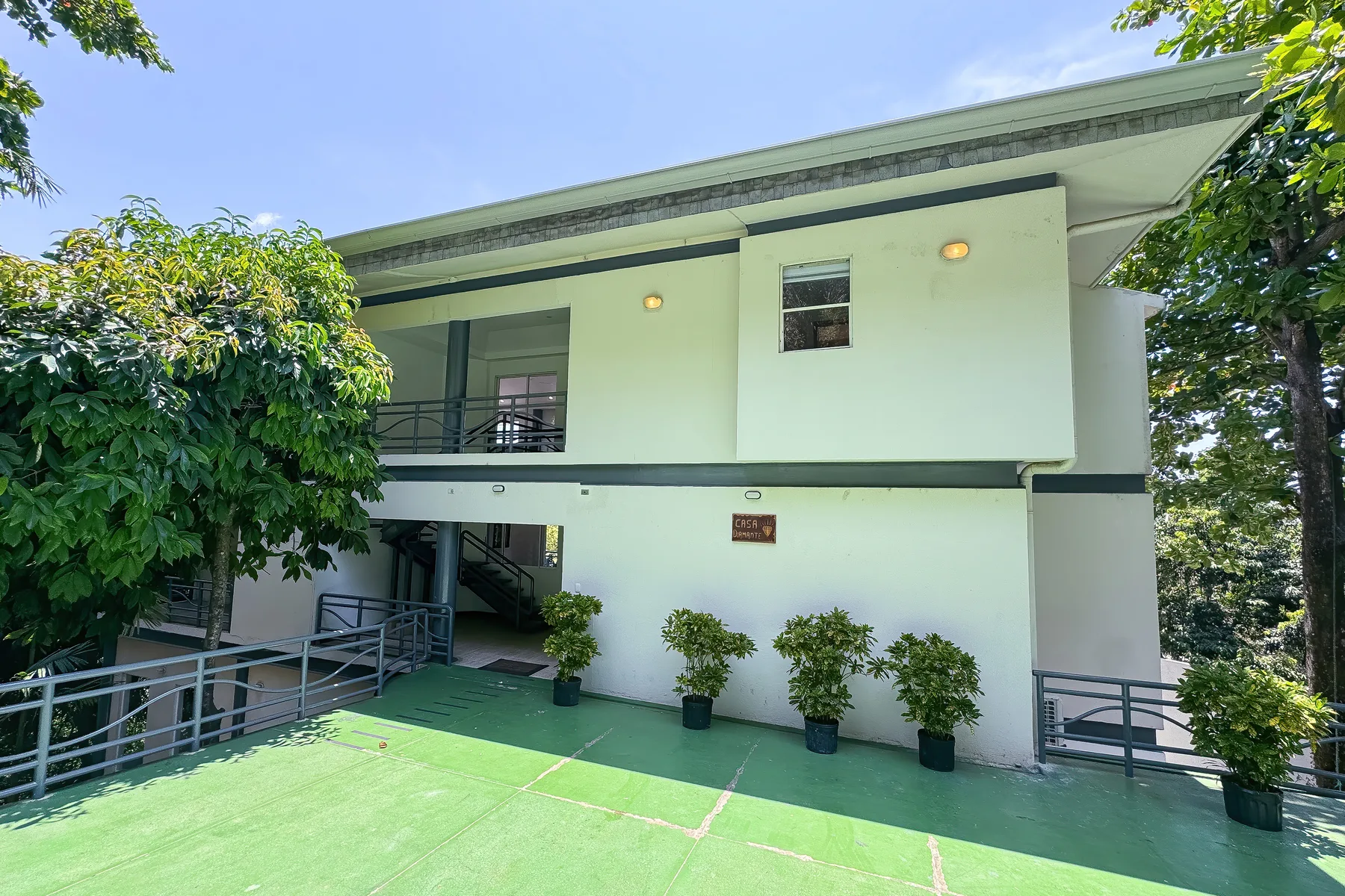 A pale green two-story building stands beside a paved outdoor area and leafy trees.