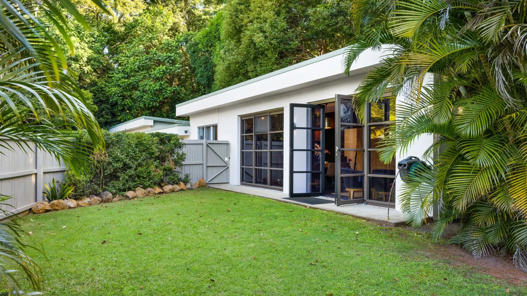 A modern white building with open glass doors faces a green lawn surrounded by tropical plants.