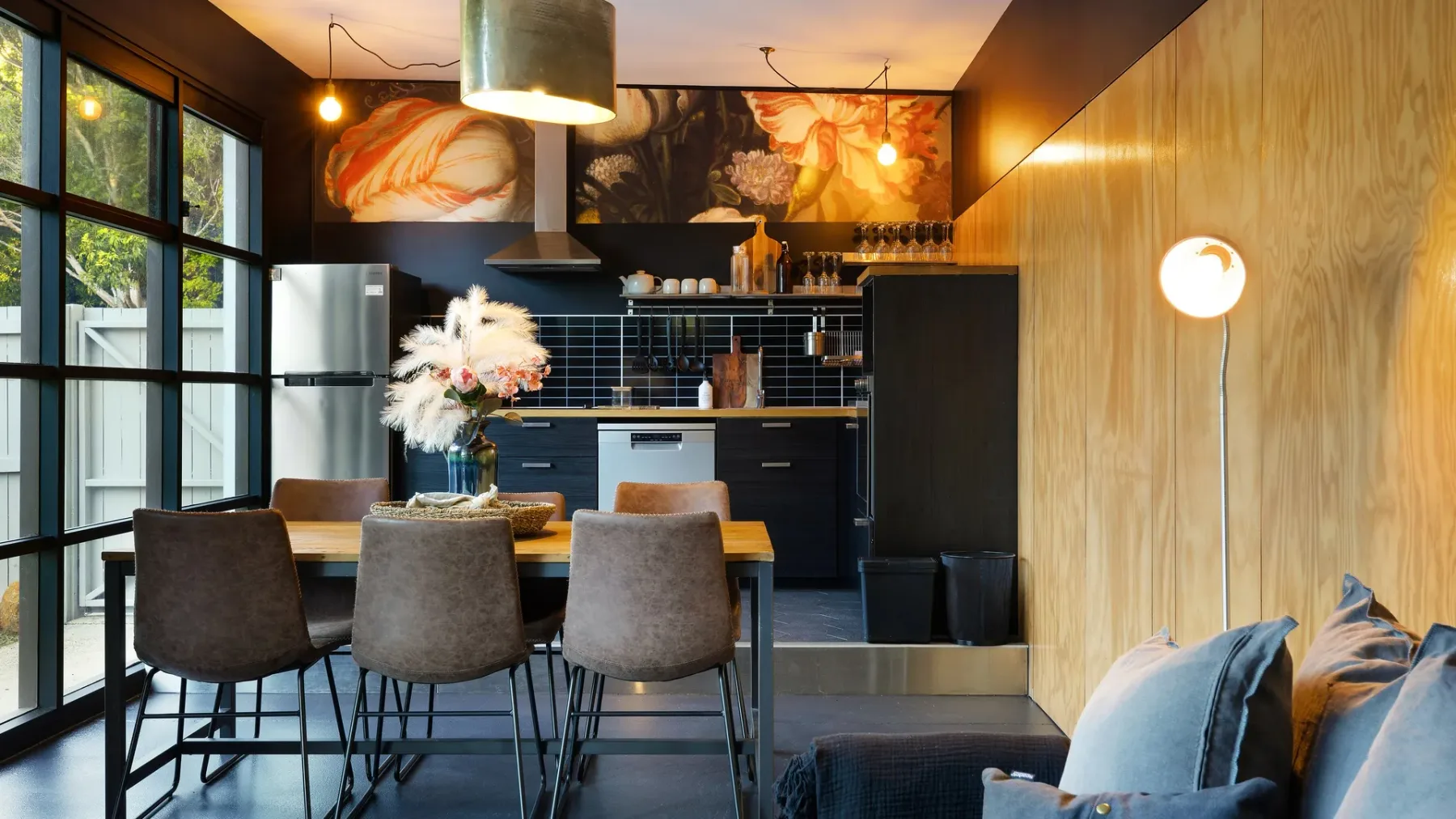 A dining area opens onto a modern kitchen, with a table beside large windows.