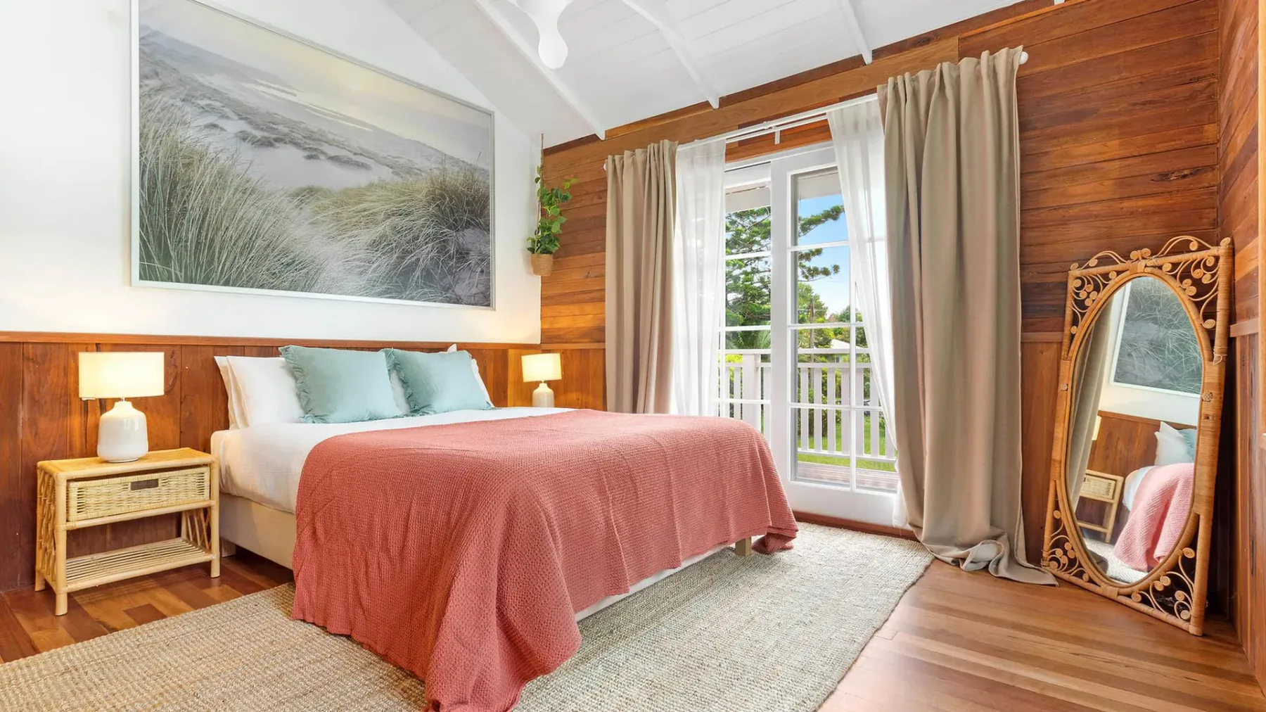 Un dormitorio con una cama cubierta por una manta coral junto a puertas de vidrio y cortinas beige.