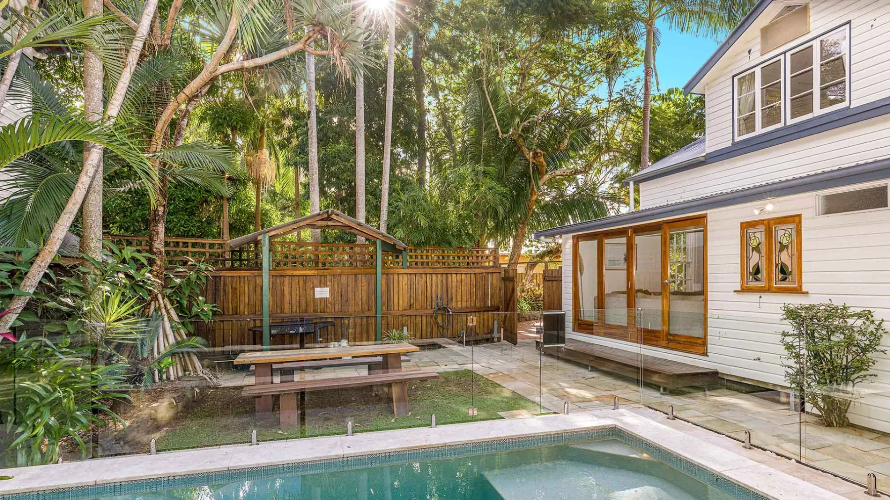 Una piscina en un patio tropical junto a una casa blanca, con una mesa de picnic bajo las palmeras.