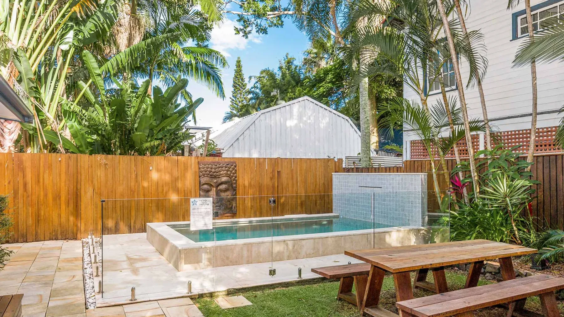 Zona de piscina al aire libre con una cerca de madera, plantas tropicales y asientos de picnic.