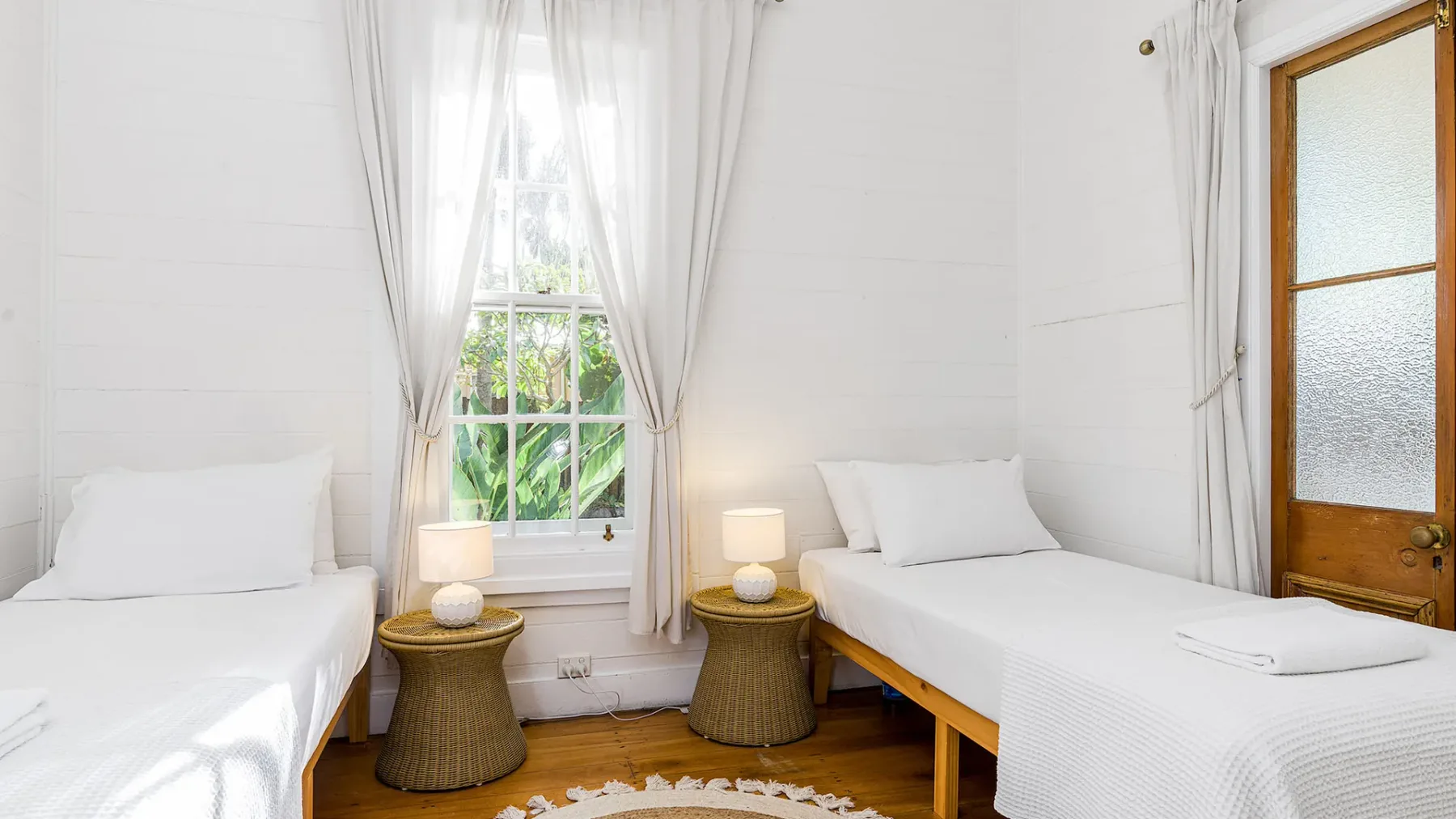 Un dormitorio luminoso con dos camas, una ventana central y una puerta de madera.