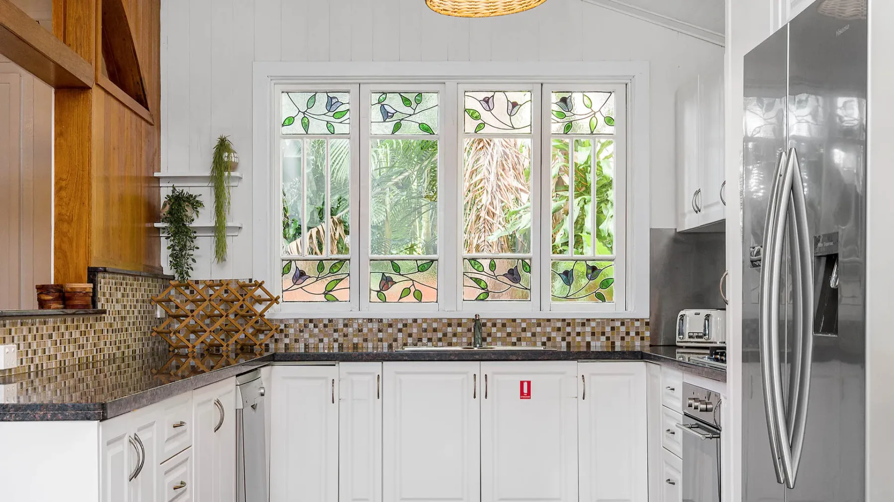 Una cocina luminosa con gabinetes blancos, ventanas decoradas y un refrigerador de acero inoxidable.