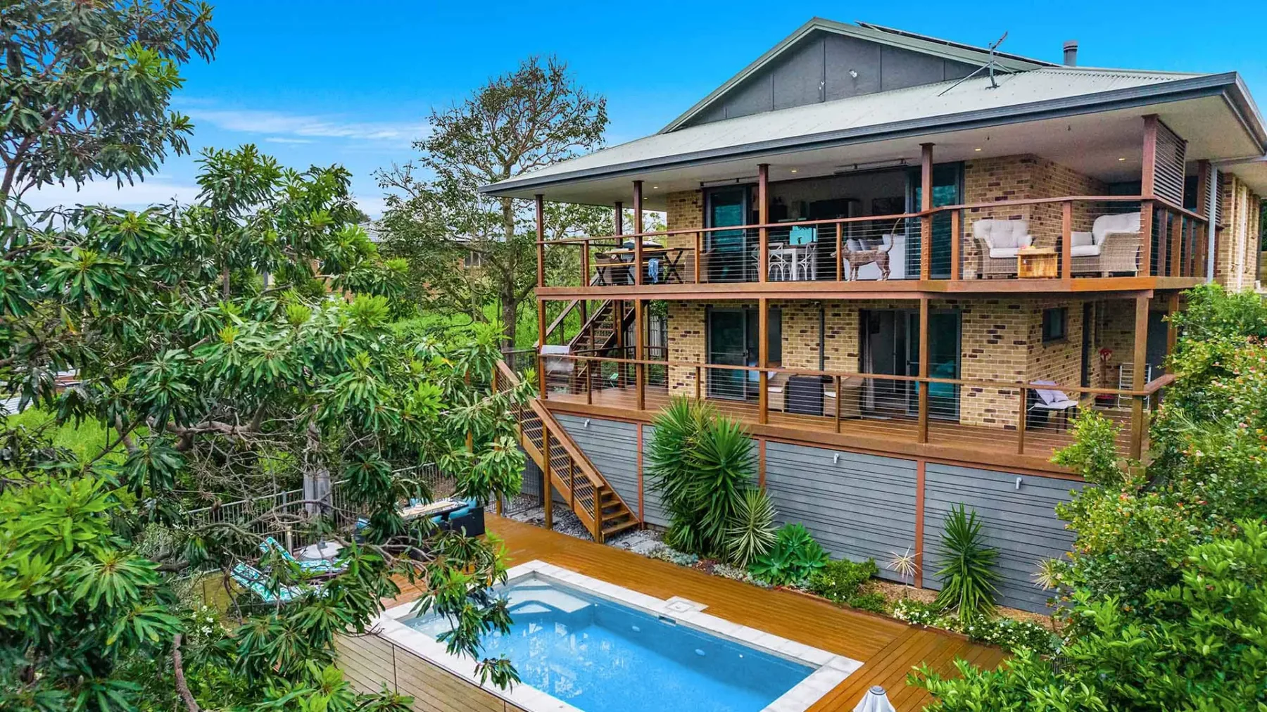 A raised house with wooden decks overlooks a swimming pool surrounded by greenery.