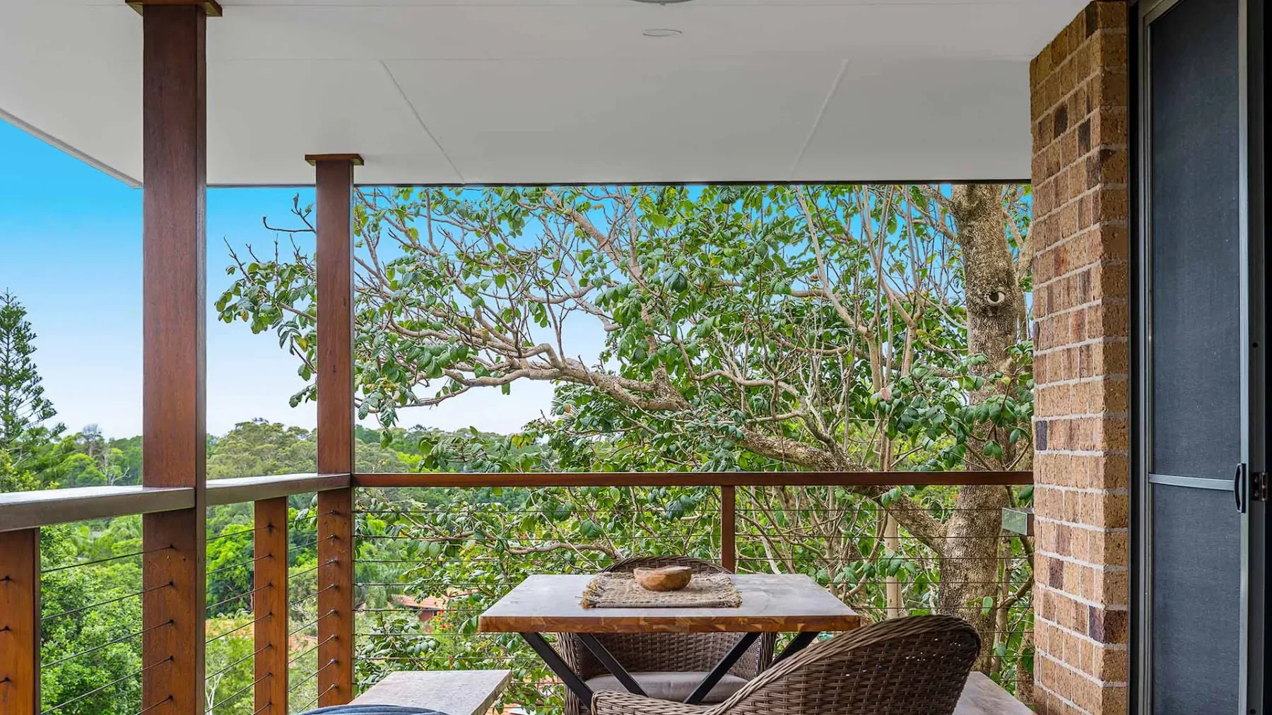 A covered outdoor balcony has a table and seating overlooking leafy trees.