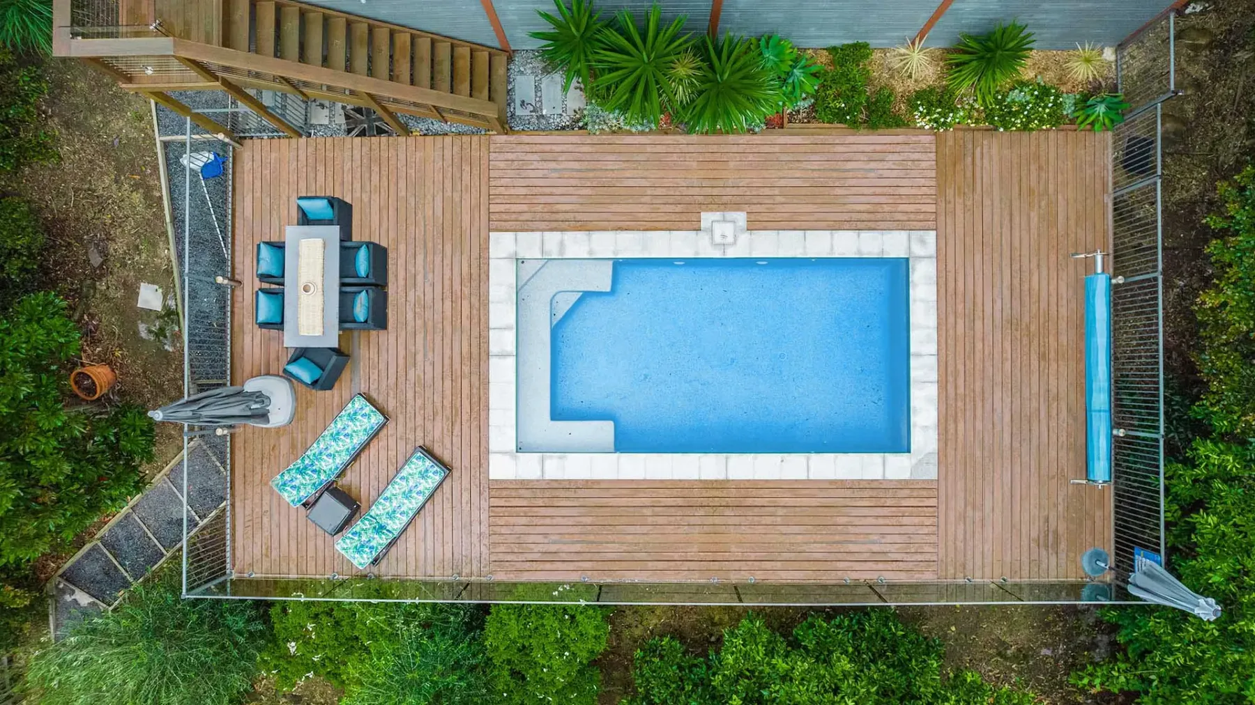 An outdoor pool sits on a wooden deck with lounge chairs and a dining table.