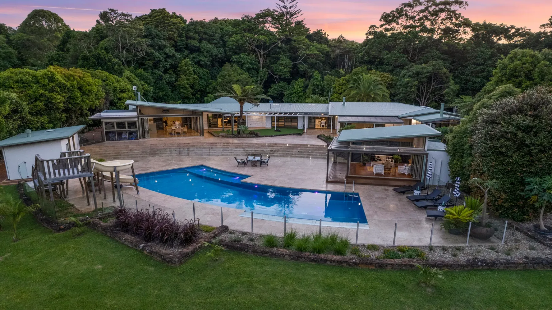 Una piscina y un amplio patio están rodeados de árboles frondosos al atardecer.