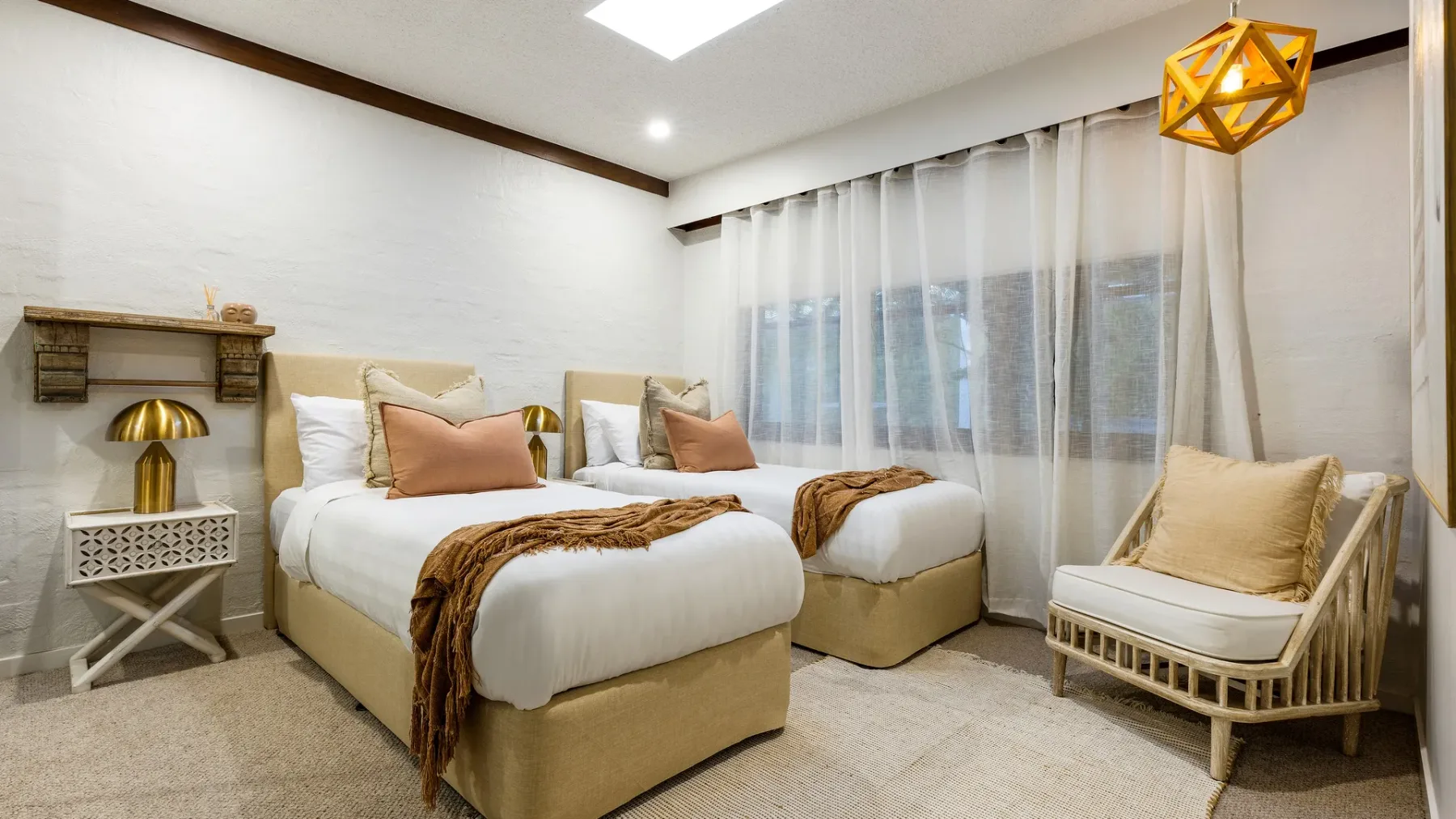 Un dormitorio con dos camas, ropa de cama en tonos neutros y cortinas translúcidas en las ventanas.