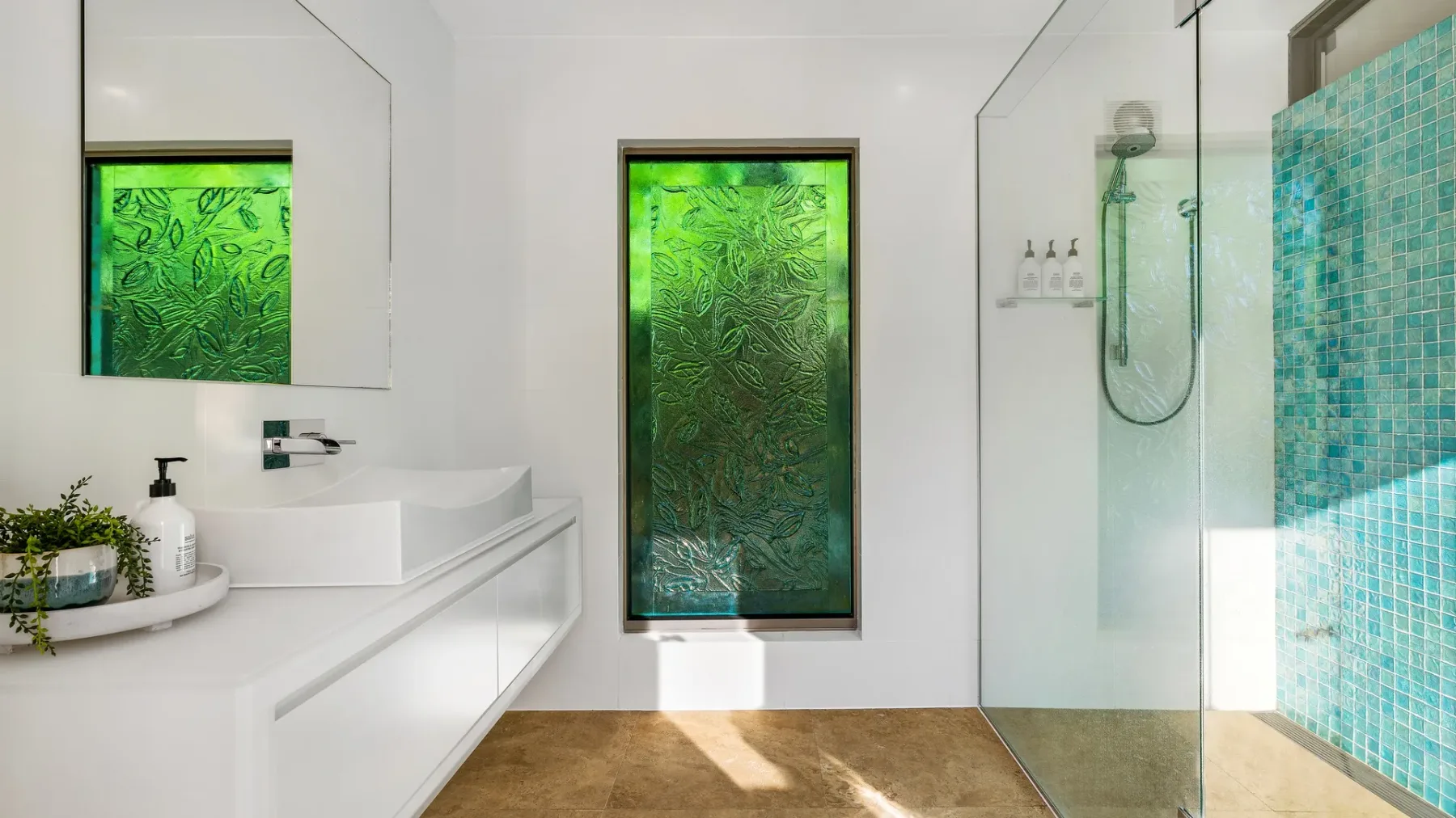 Un baño luminoso con lavabo blanco, ducha de vidrio y ventanas con motivos verdes.