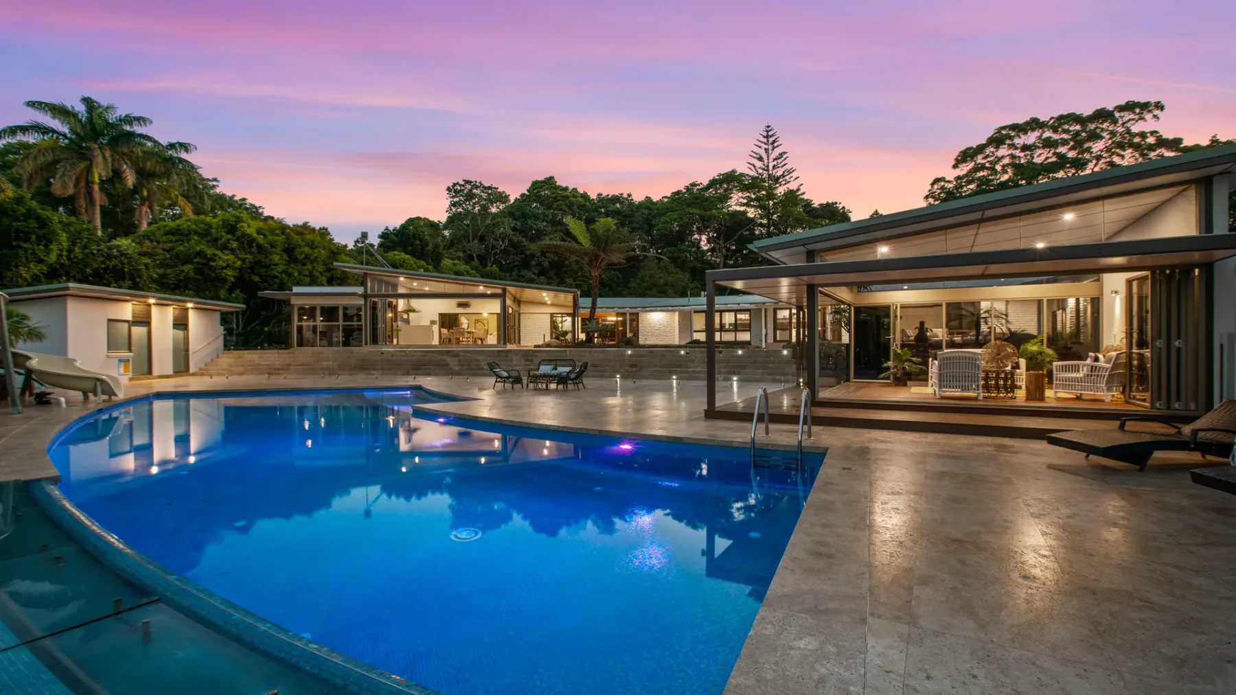Una casa moderna se abre a una piscina iluminada bajo un cielo colorido al atardecer.