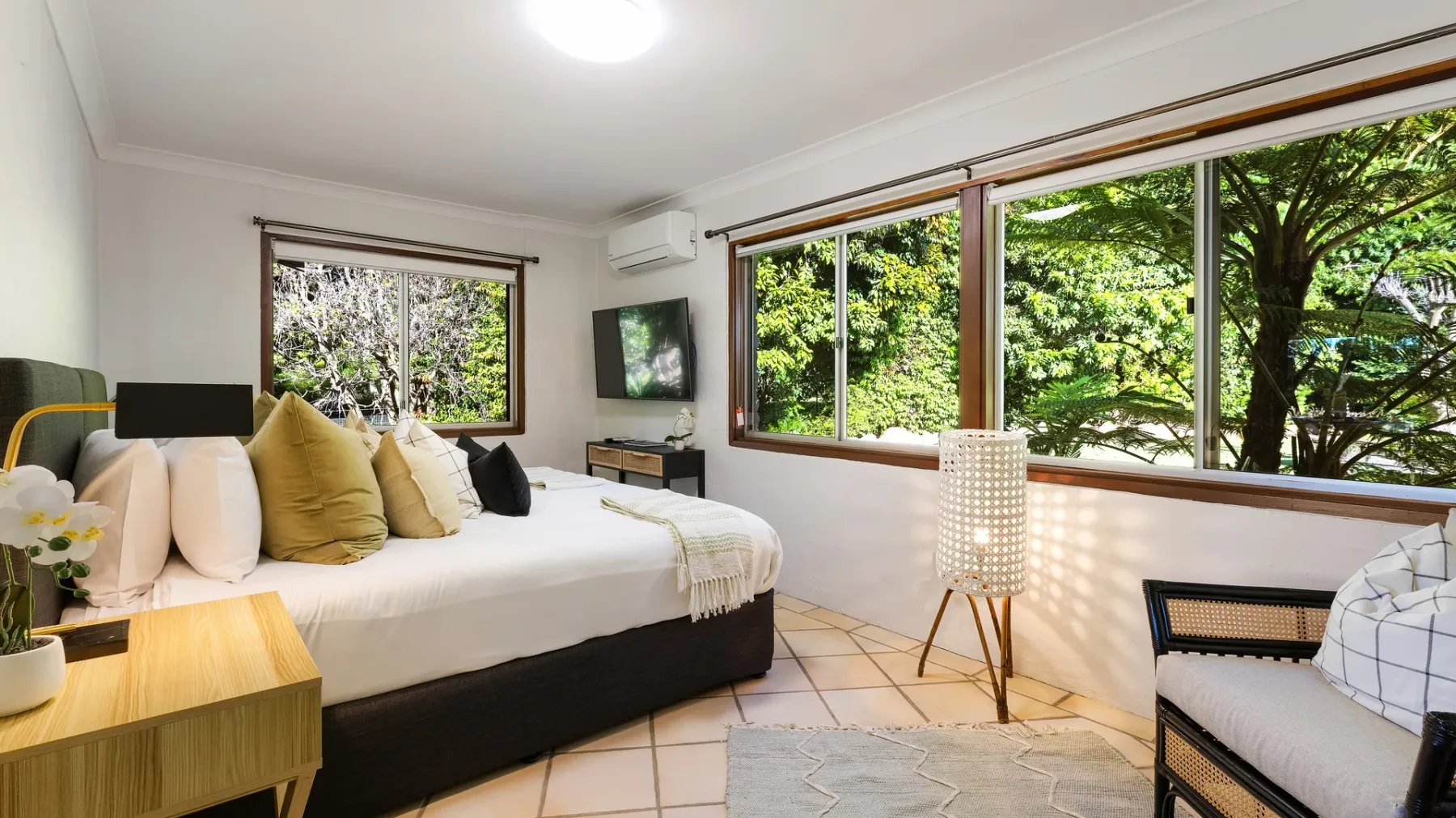 Un dormitorio luminoso con una cama grande, una mesita y ventanas con vistas a la vegetación.