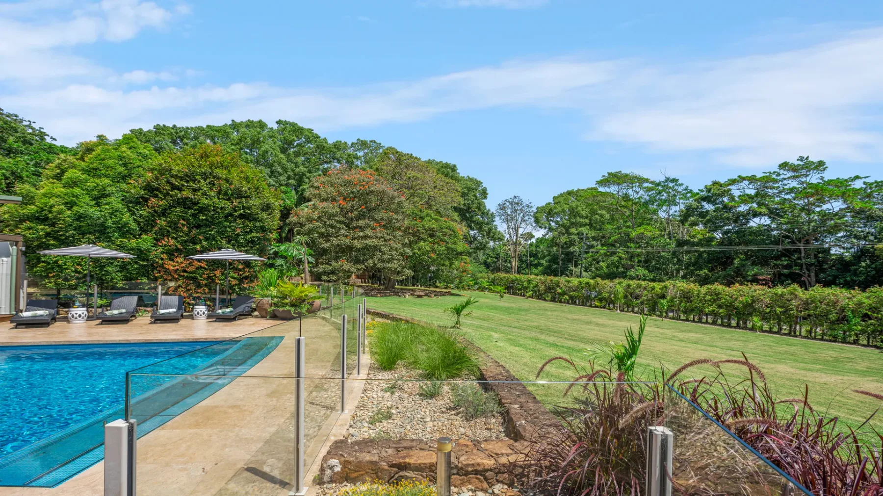 Una piscina exterior limita con un jardín y una zona de césped bajo un cielo azul.