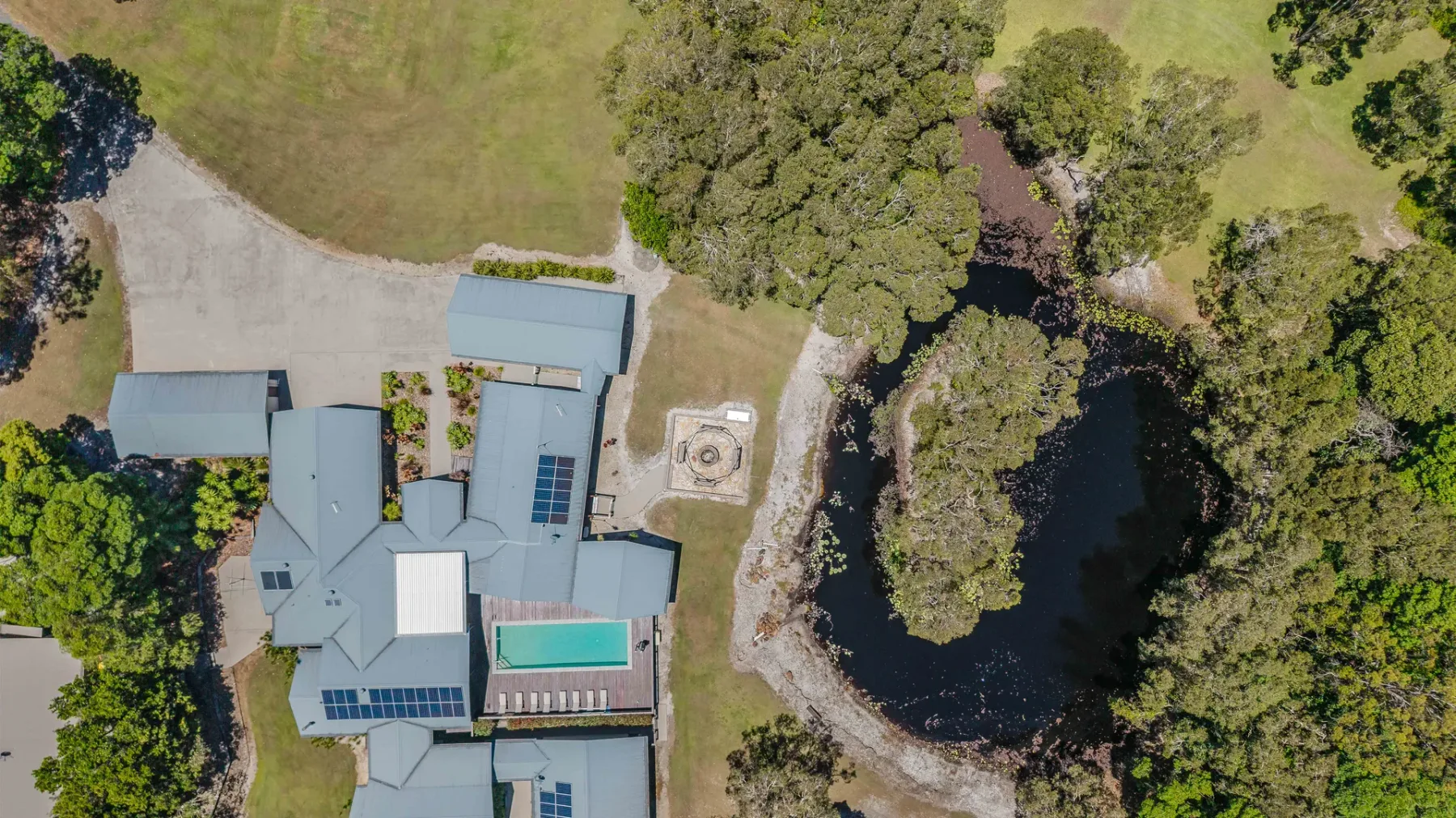 Aerial view of a building beside a pond, surrounded by trees and open lawn.