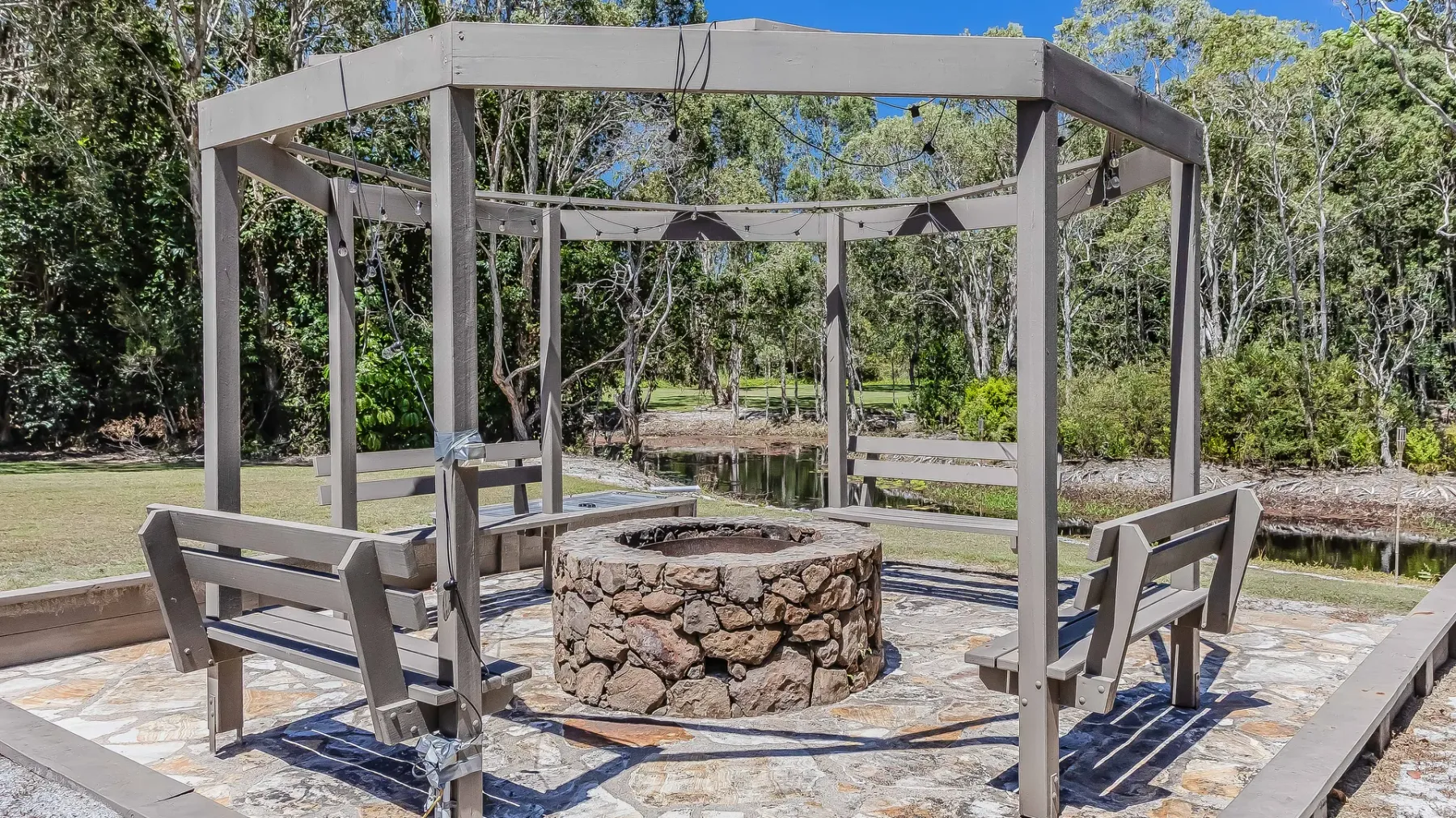 An outdoor seating area surrounds a circular stone fire pit beneath a wooden pergola.