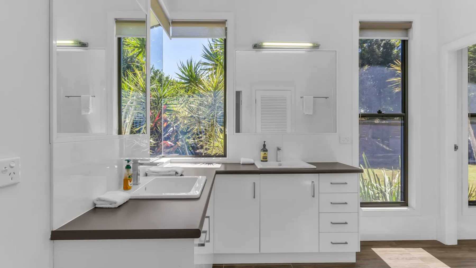 A bright bathroom vanity with a sink, white cabinets, and windows overlooking greenery.