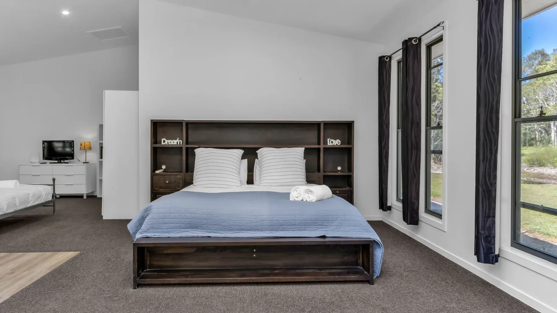 A bedroom with a bed beneath a dark shelving headboard and tall windows.