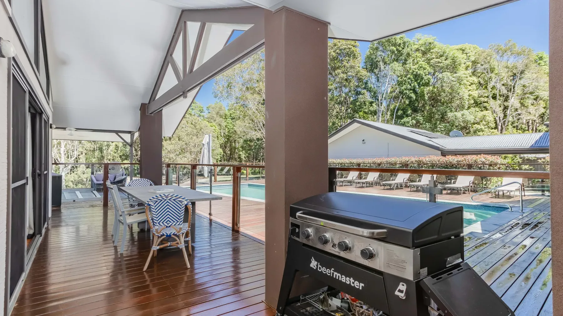 A covered outdoor deck overlooks a swimming pool, with a grill beside the seating.
