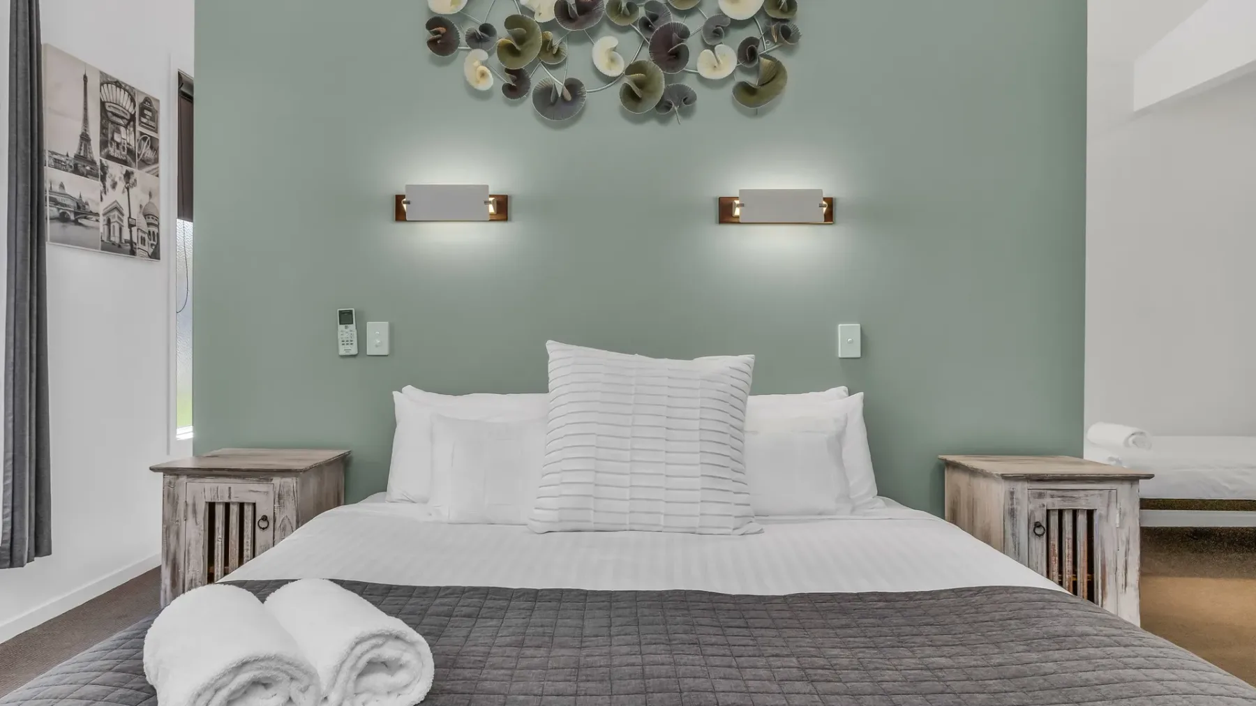 A neatly made bed sits against a pale green wall with decorative accents.
