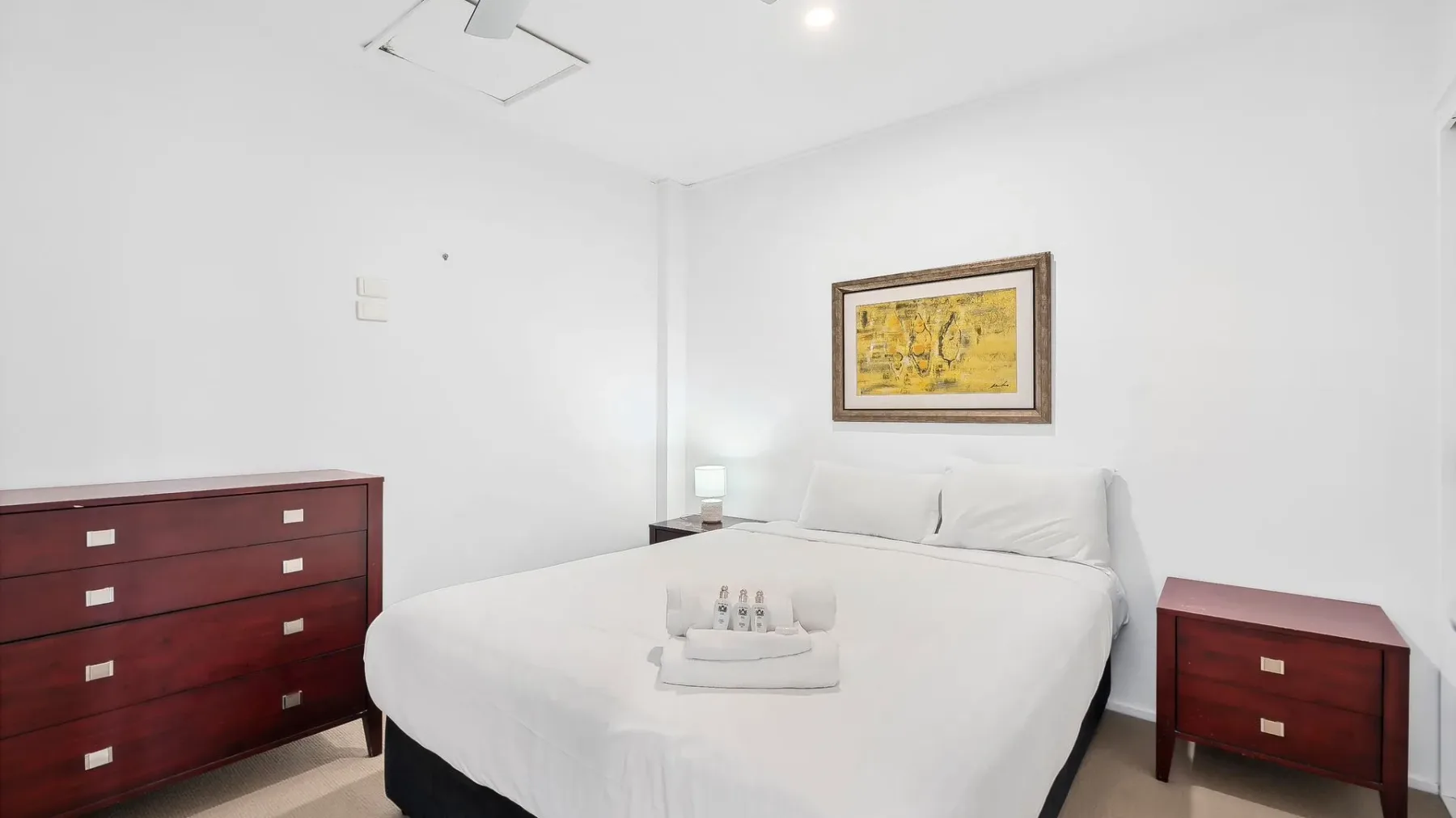 Un dormitorio con una cama tendida, cómodas rojas y un cuadro enmarcado.