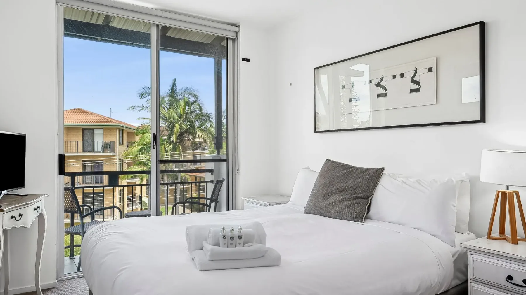 Un dormitorio luminoso con ropa de cama blanca, un cojín gris y puertas corredizas de vidrio.