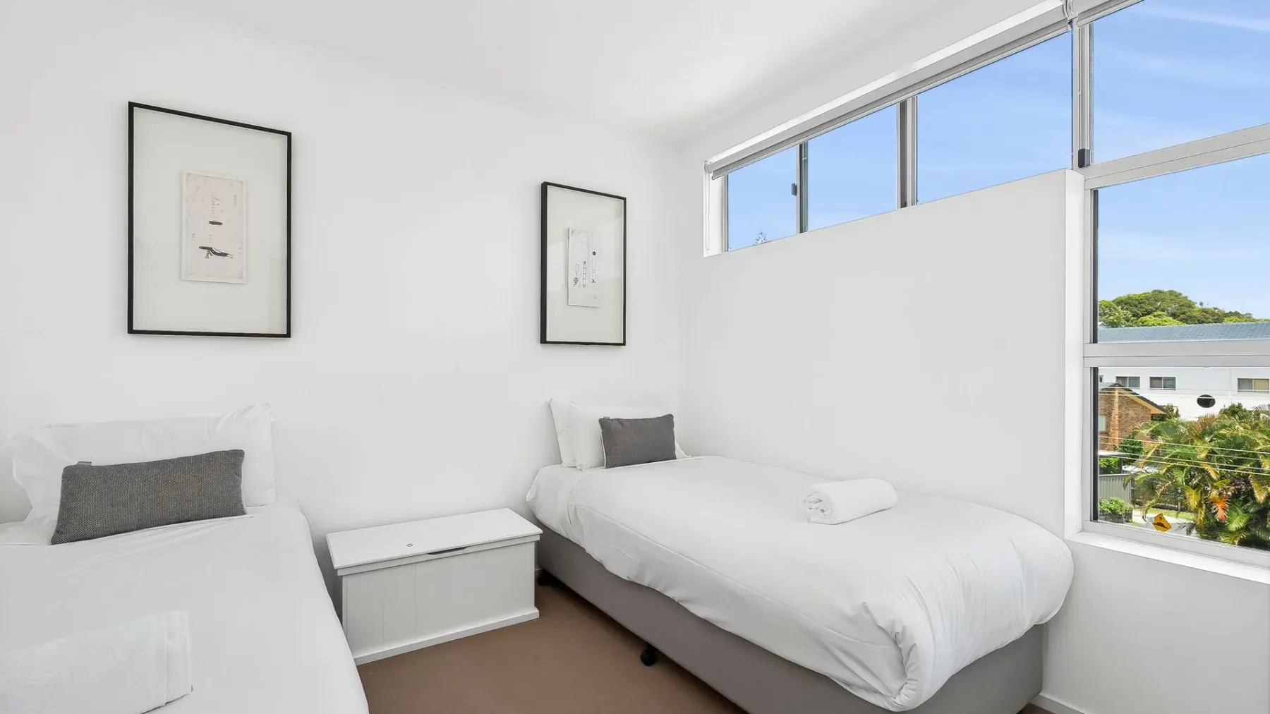 Un dormitorio luminoso con dos camas, cuadros y ventanas grandes.