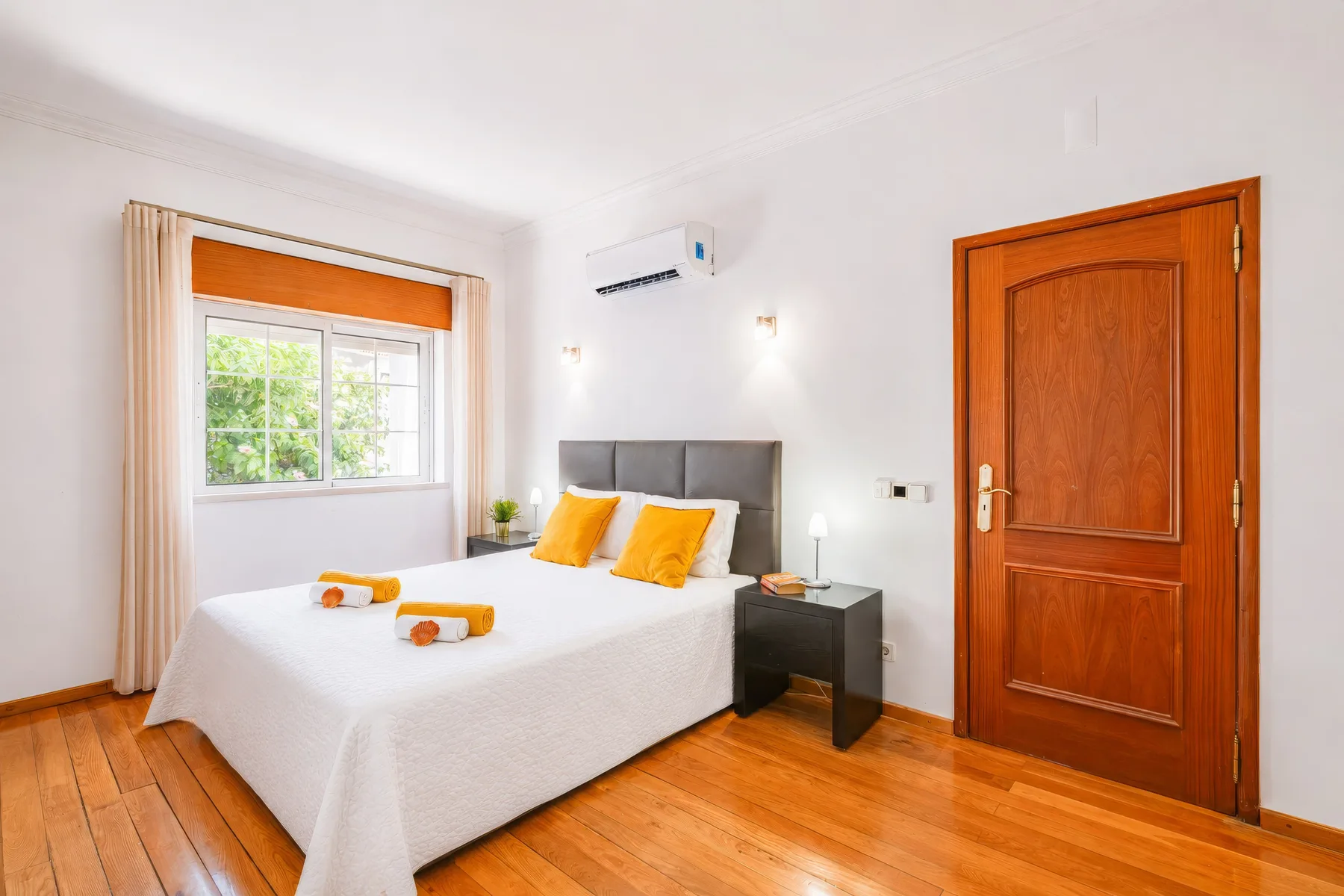 Un dormitorio con una cama, cojines naranjas, una ventana y una puerta de madera.