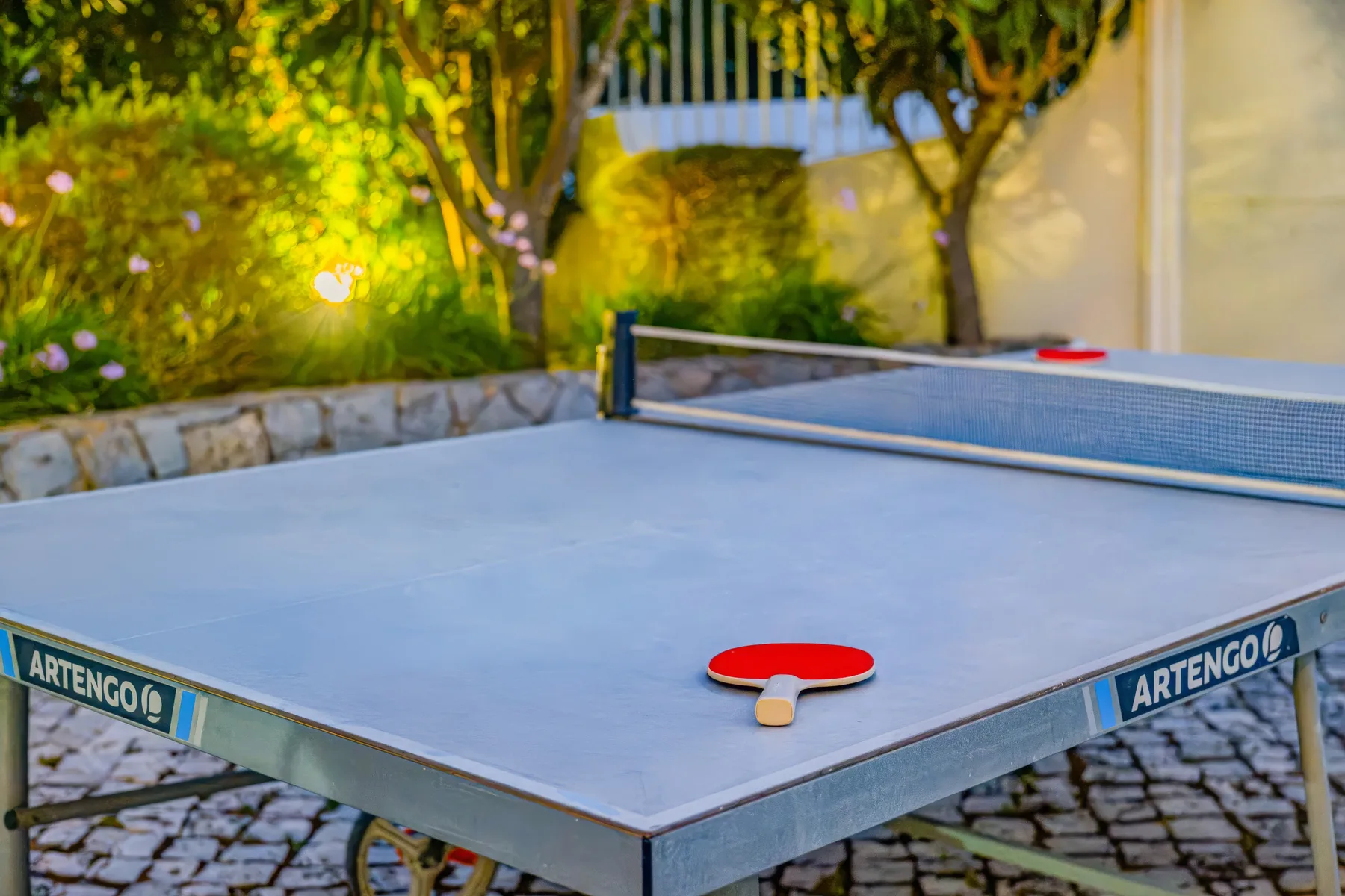 Una mesa de ping-pong al aire libre con palas está junto a un jardín frondoso.