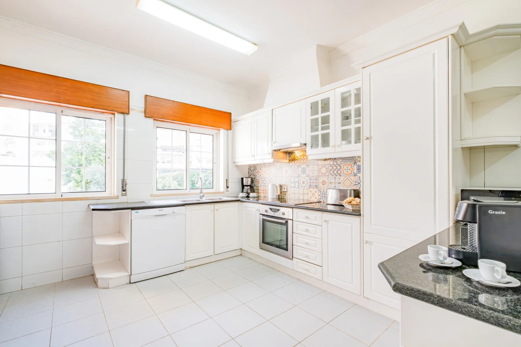 Una cocina luminosa con armarios blancos, suelo de baldosas y ventanas sobre las encimeras.