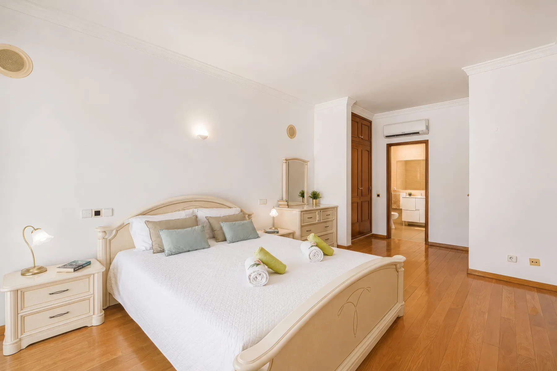 Un dormitorio luminoso con una cama hecha, mesitas de noche y una puerta que da al baño.