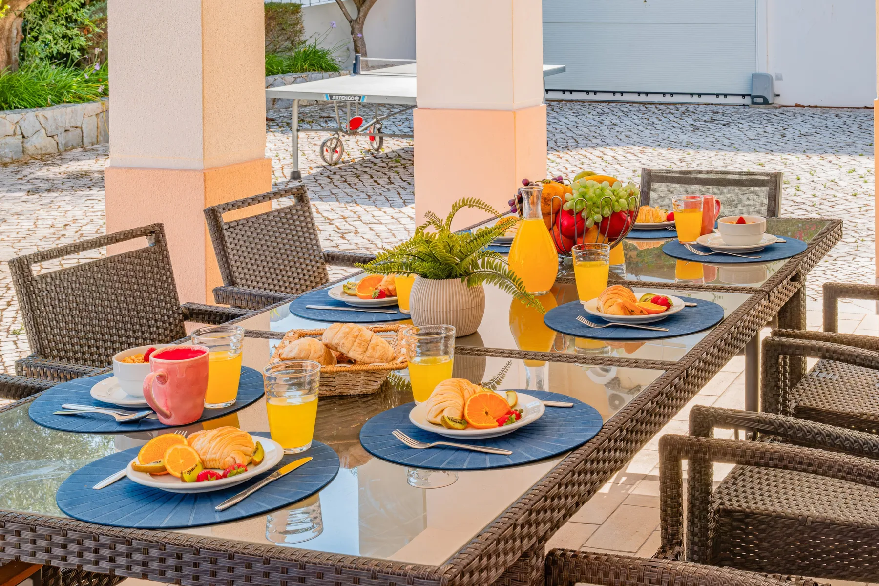 Una mesa de comedor al aire libre está puesta con platos, bebidas, bollería y fruta.