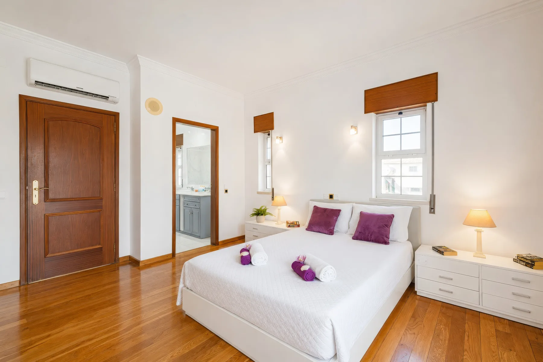 Un dormitorio luminoso con una cama blanca, cojines morados y suelo de madera.