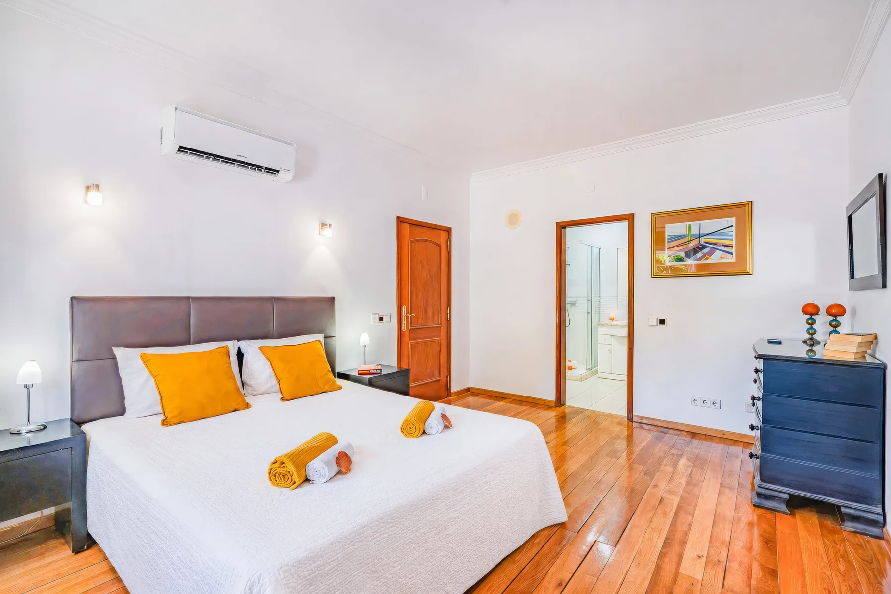 Un dormitorio luminoso con una cama, suelo de madera y una puerta hacia el baño.