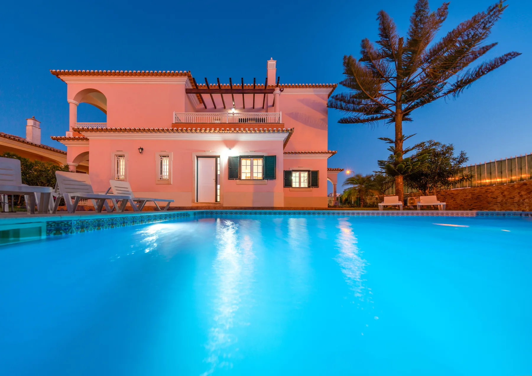 Una piscina iluminada frente a una casa rosa al anochecer.
