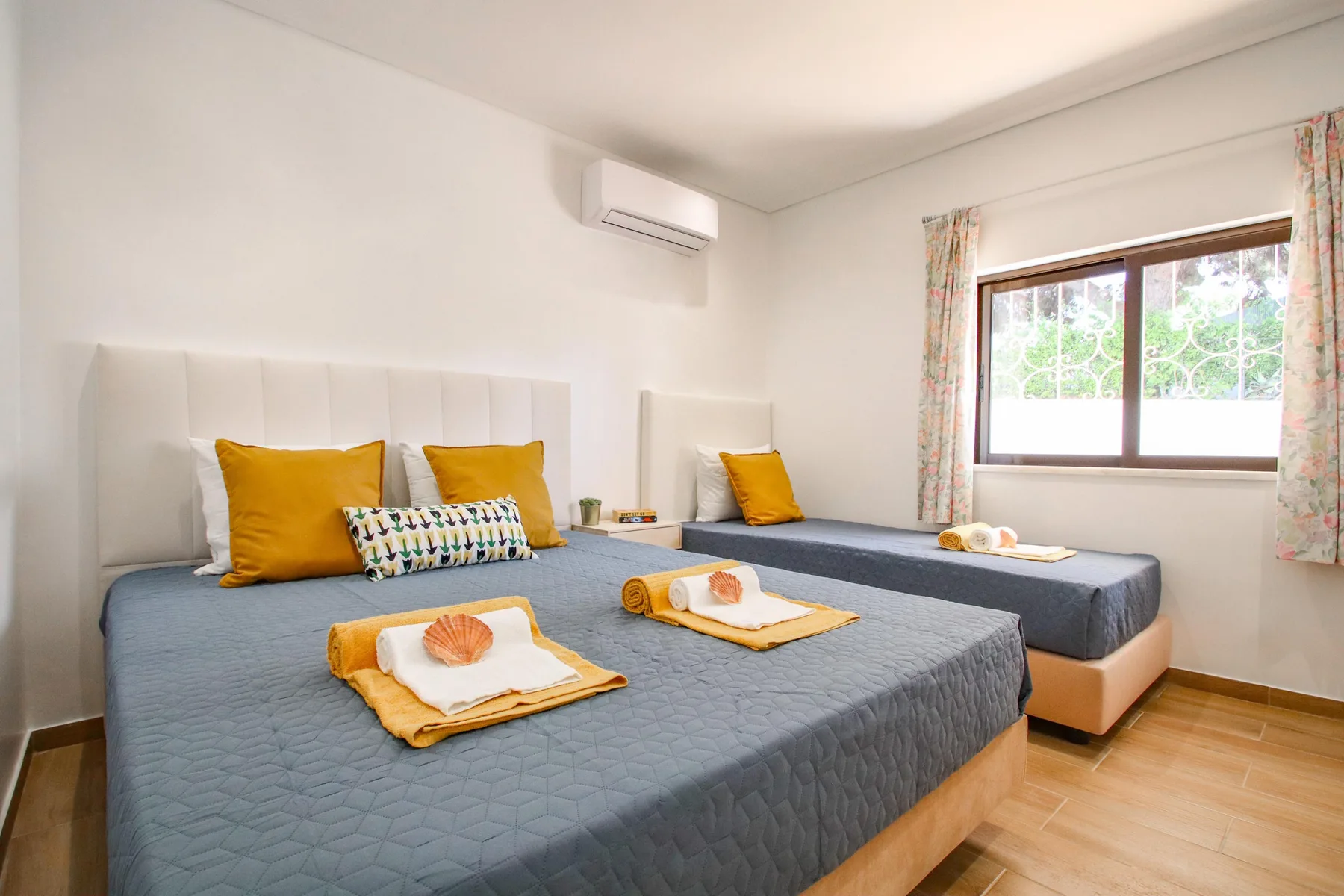 Un dormitorio luminoso con dos camas, cojines de colores y una ventana con cortinas estampadas.