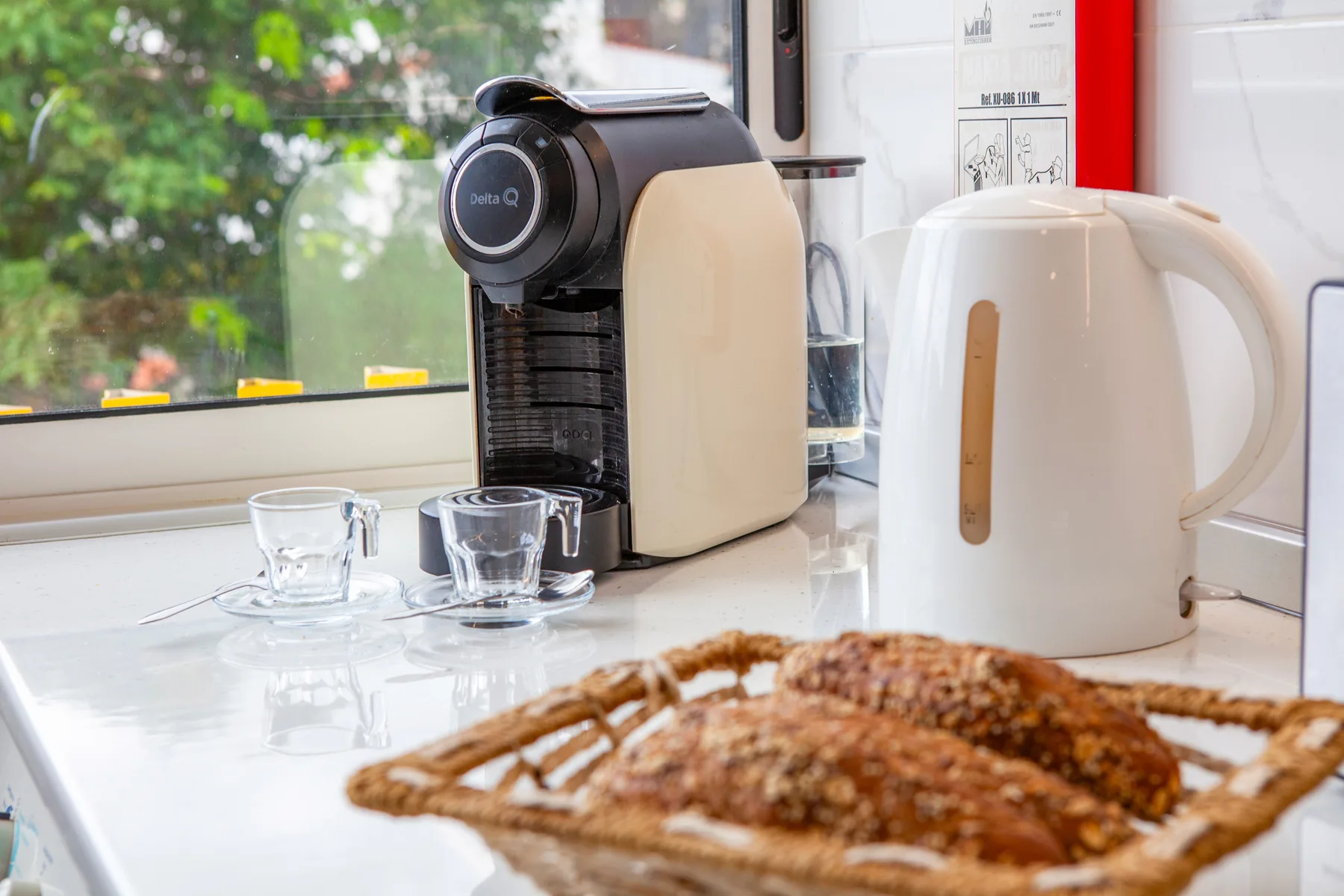 A countertop coffee maker and electric kettle sit beside cups by a window.