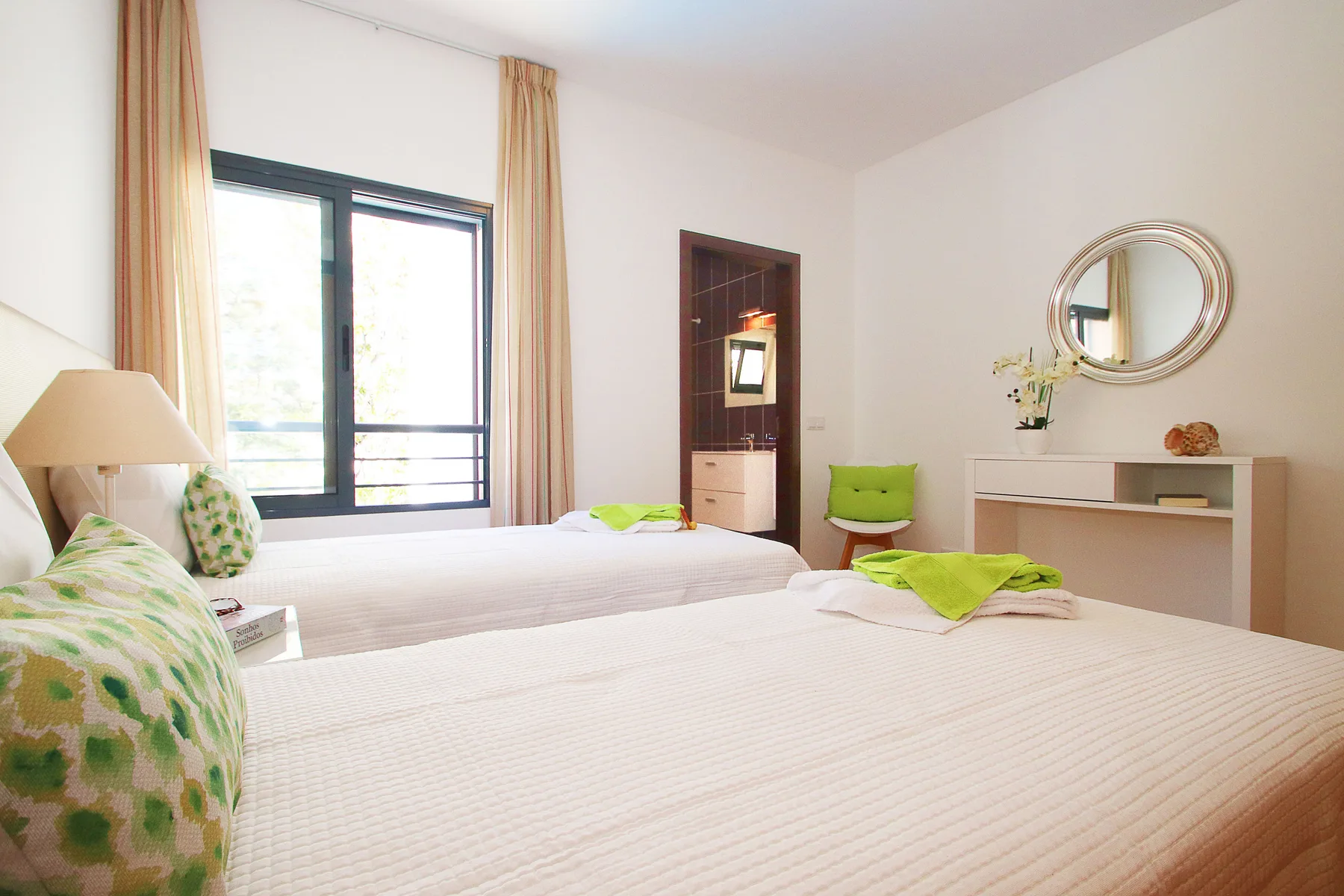 Un dormitorio luminoso con dos camas, una ventana grande y un espejo redondo.