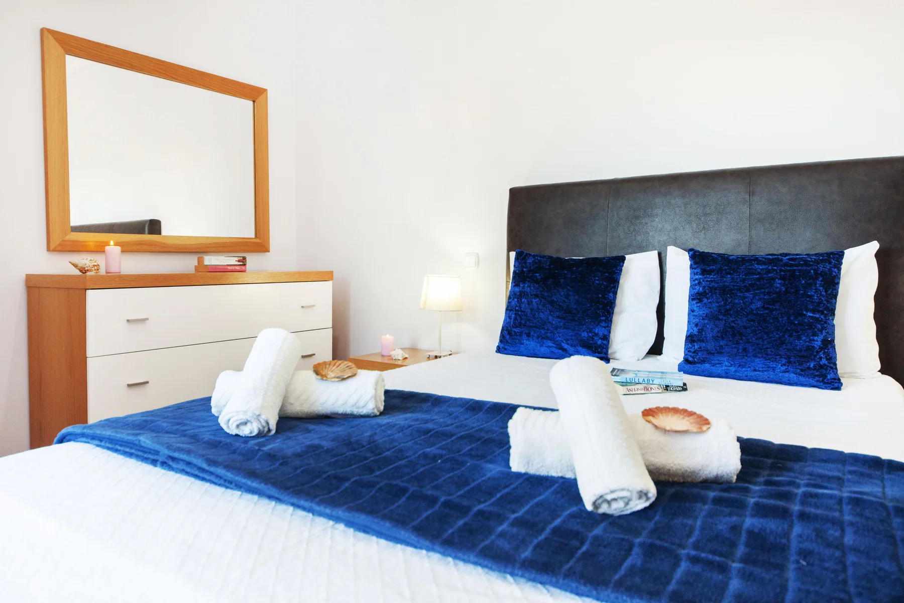 A bedroom with blue bedding, white pillows, and a dresser beside the bed.