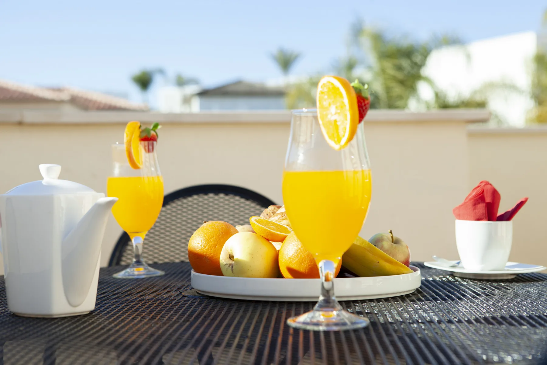 A sunny outdoor table with orange juice, citrus fruit, and a white teapot.