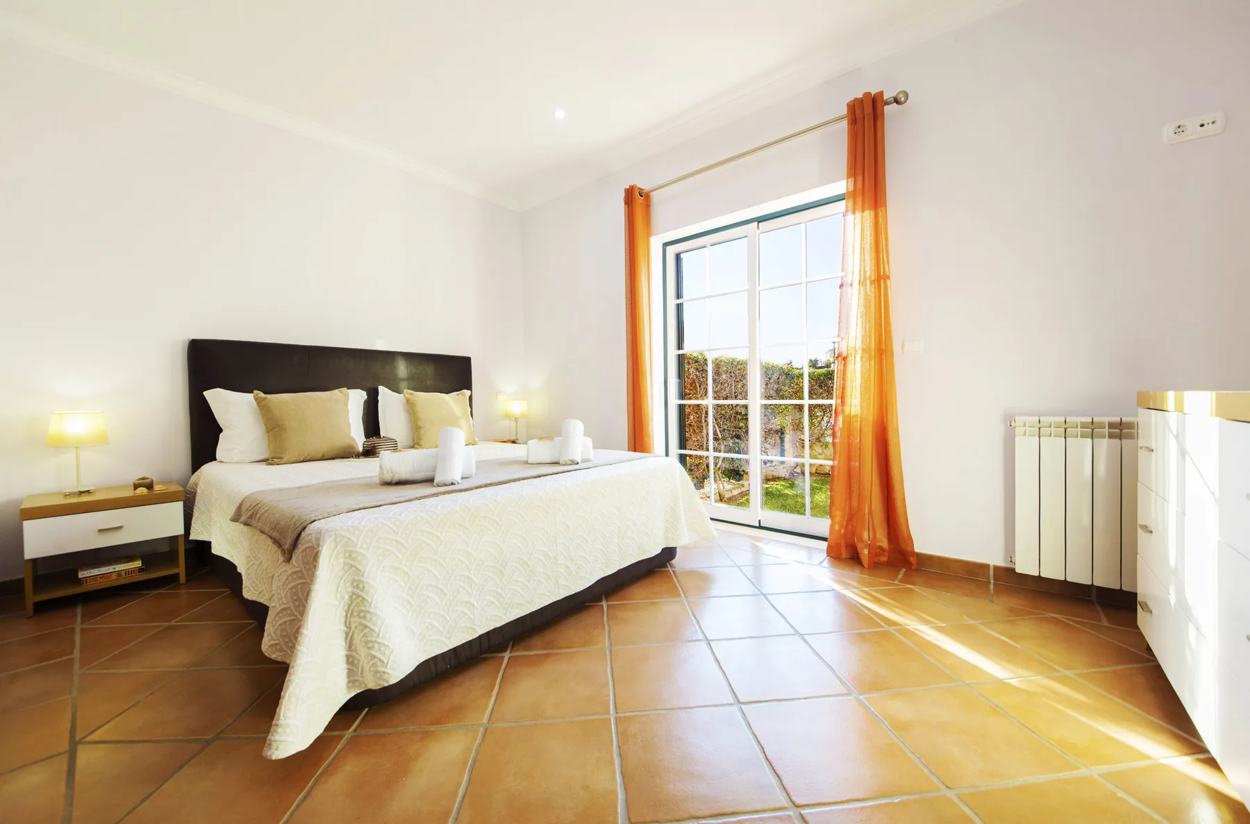 A bright bedroom with a bed beside a large window framed by orange curtains.