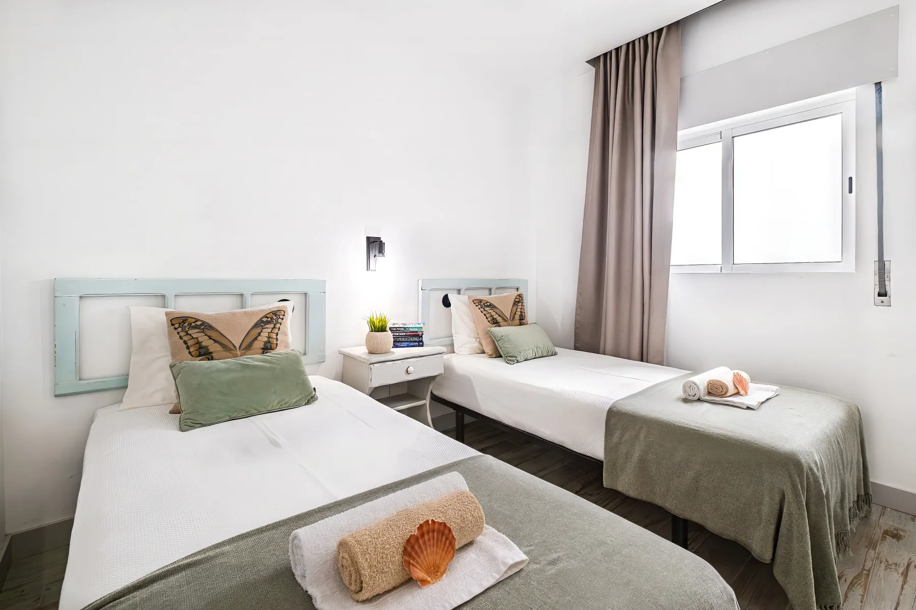 A bright bedroom with two beds, pale bedding, and a window with curtains.