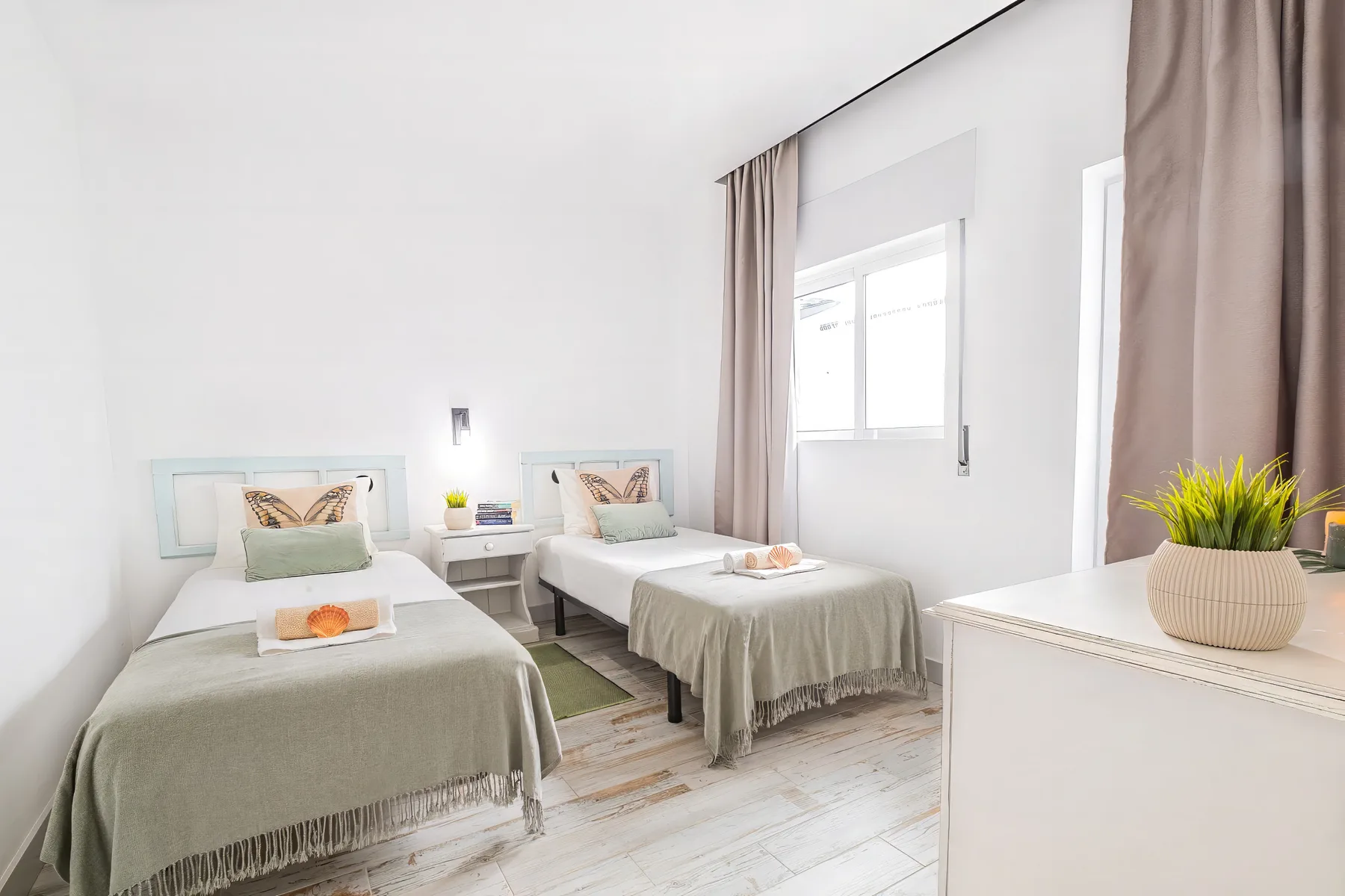 A bright bedroom with two beds, pale bedding, and a window with curtains.