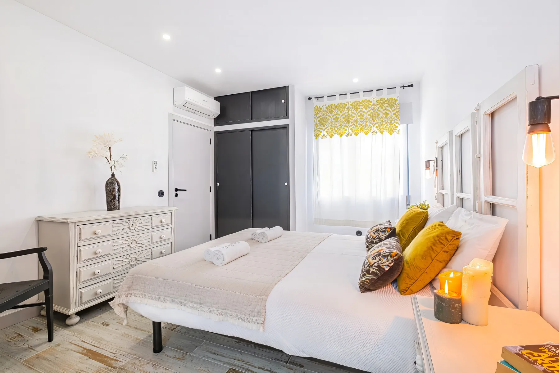 A bright bedroom with white bedding, mustard cushions, and a dresser.