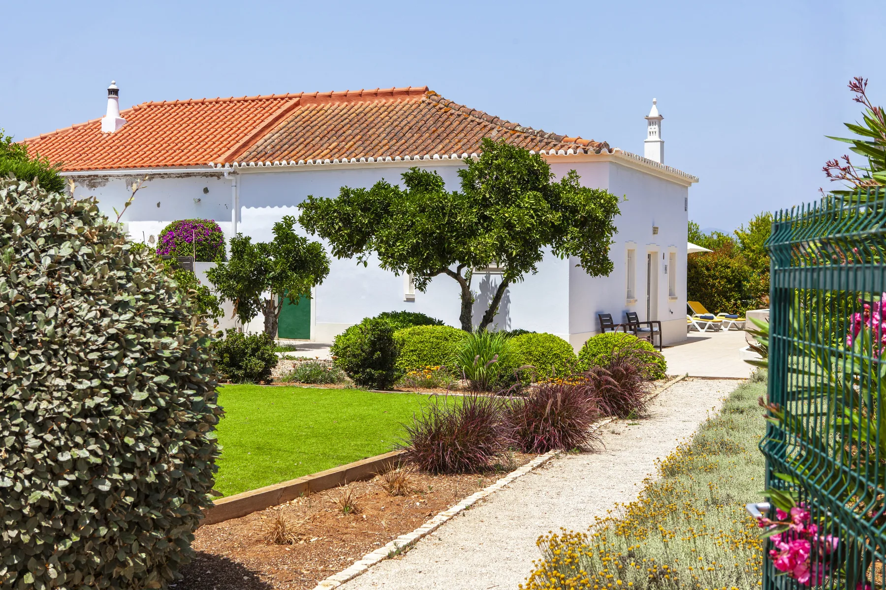 A white house with a tiled roof stands beside a garden and gravel path.