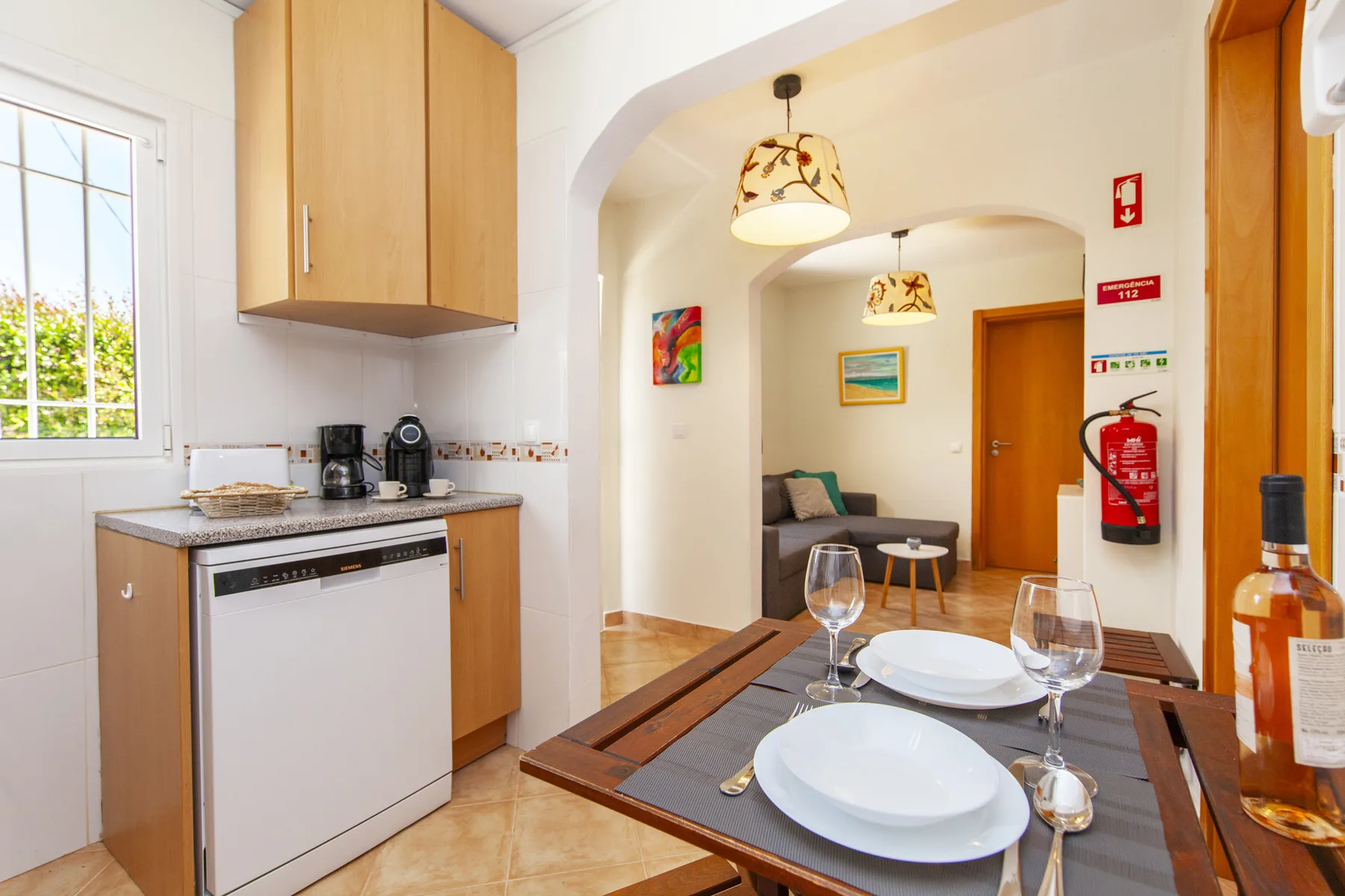 A dining table set with plates sits beside a compact kitchen with wooden cabinets.