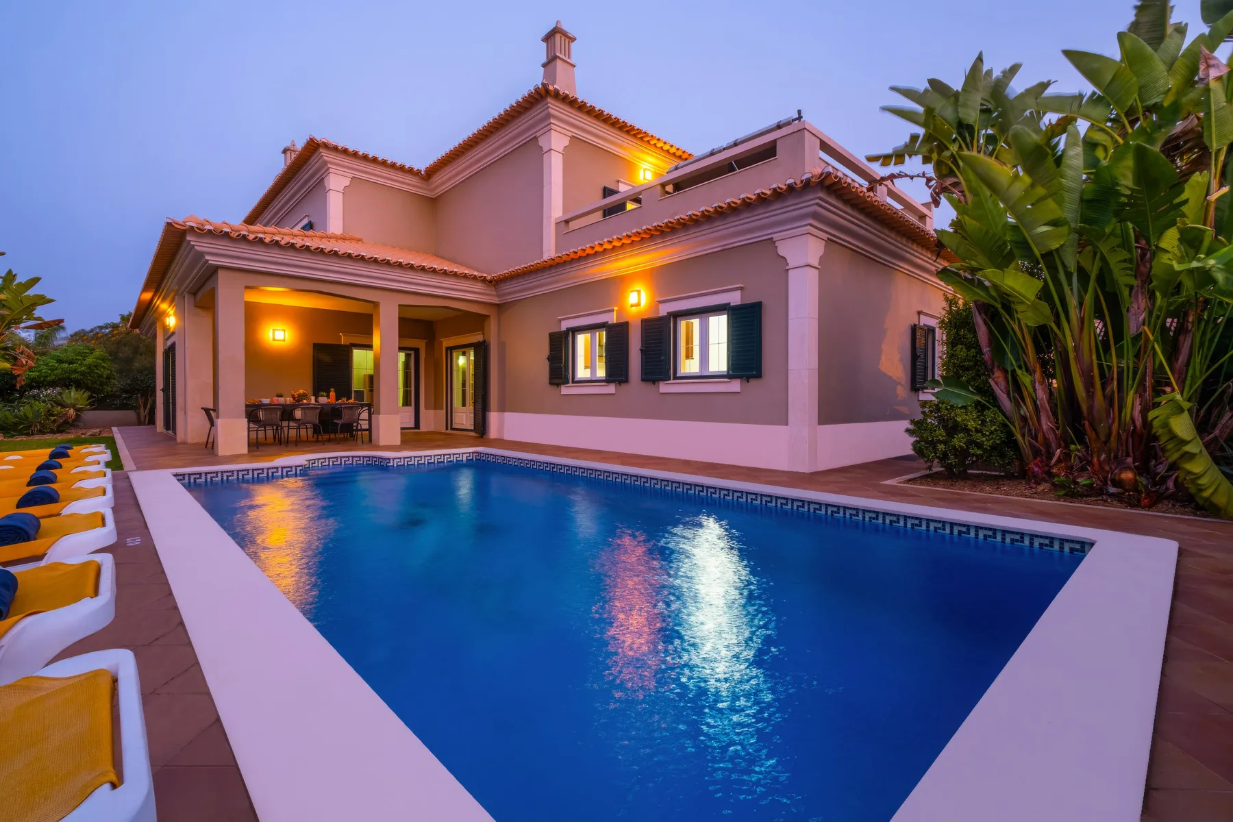 Una piscina junto a una villa rosa, con un patio cubierto y plantas tropicales.