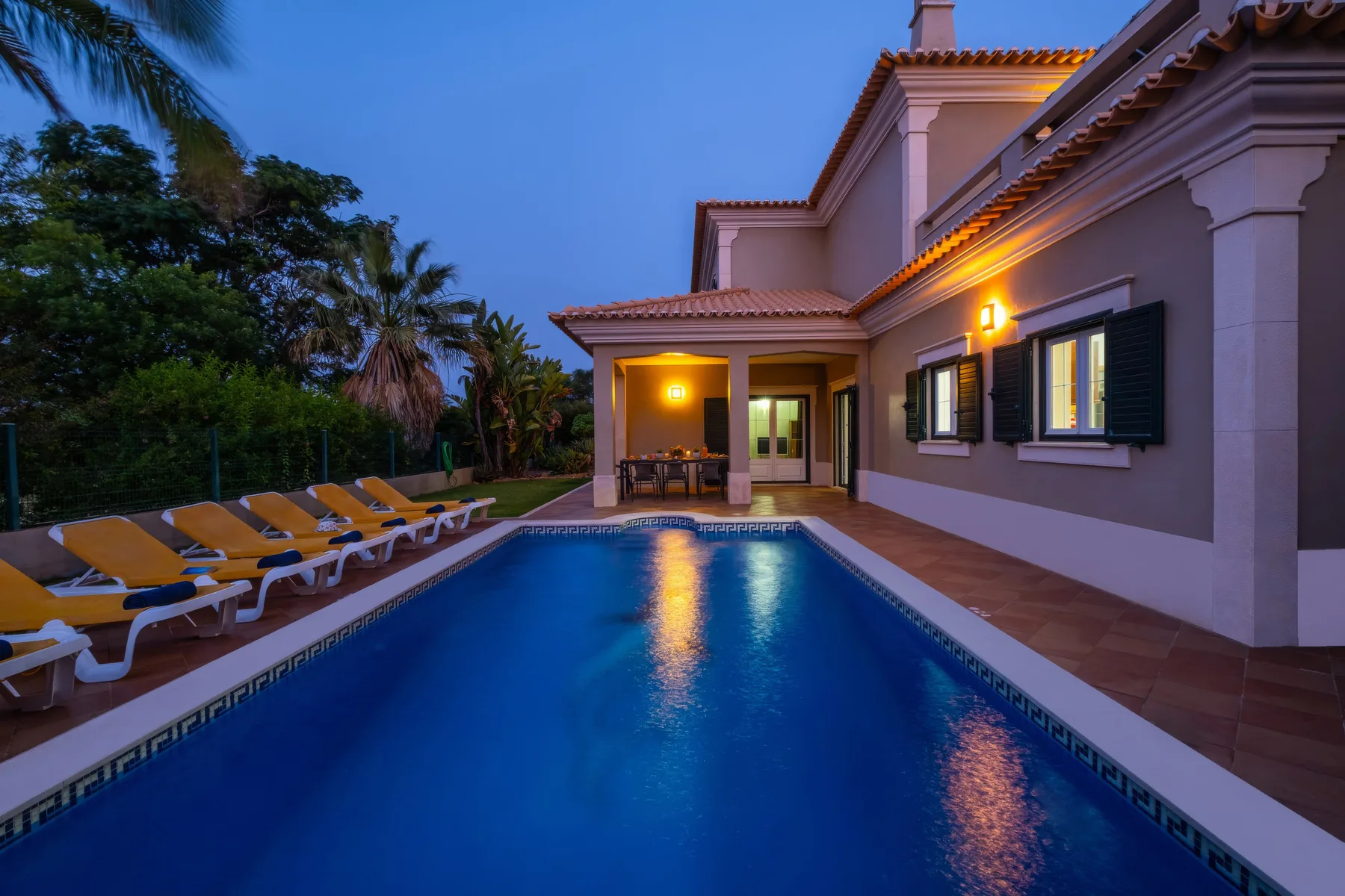 Una piscina junto a una casa, con asientos de descanso a lo largo del borde.